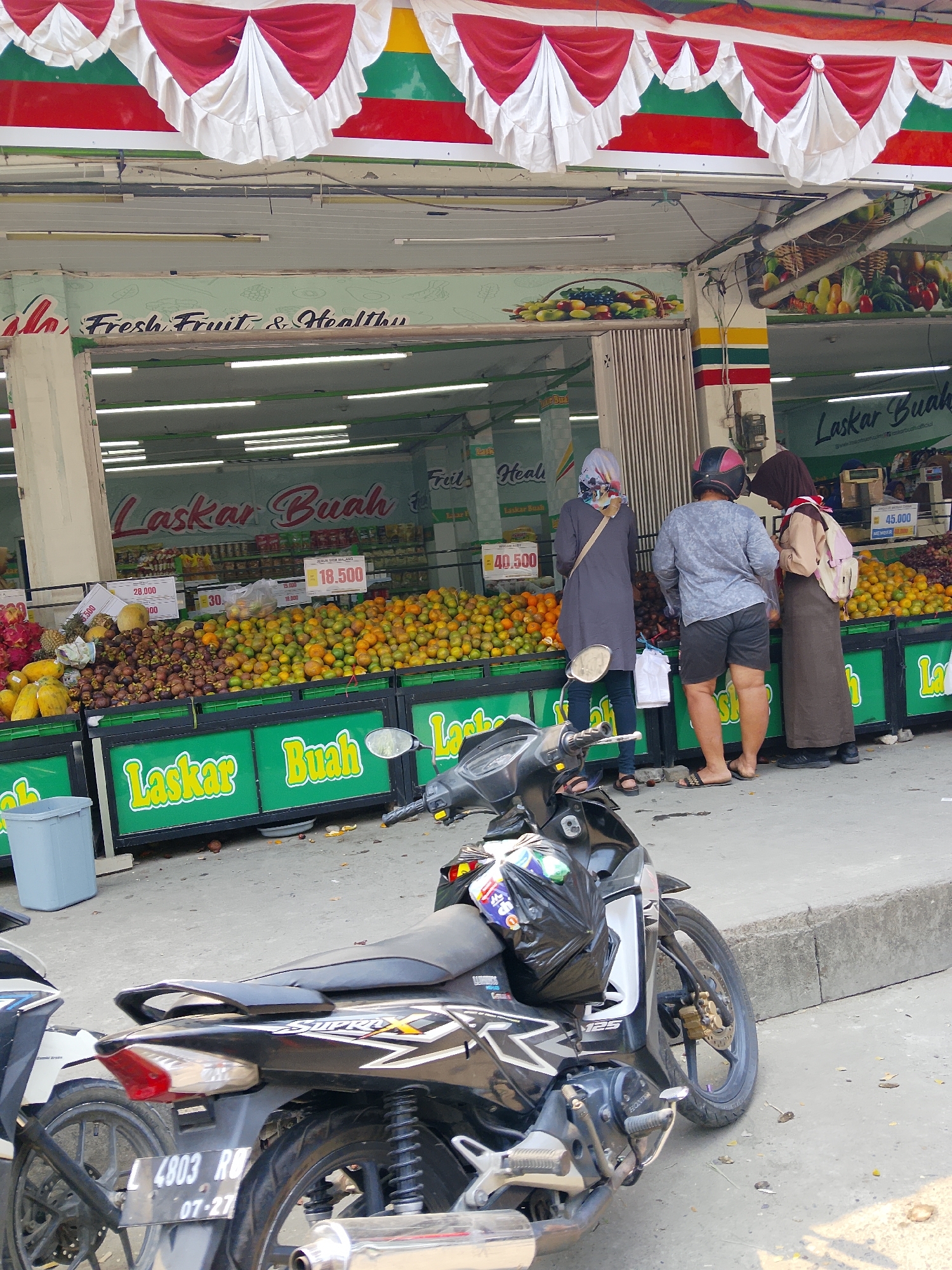 The image depicts an outdoor fruit market in Karangboyo, Indonesia. In the foreground, a black motorcycle is parked. Behind it, a row of green stalls displays a large variety of fruits, including oranges, mangosteens, papayas, and dragon fruit. Three people are visible in front of the stalls, appearing to shop. One person, wearing a grey top and black shorts, is facing away from the camera, while two others, dressed in hijabs and modest clothing, are interacting with the fruit displays. The stalls have signs with Indonesian text and prices. In the background, the market structure is visible with colorful awnings and signage. The overall scene is characteristic of a local marketplace.