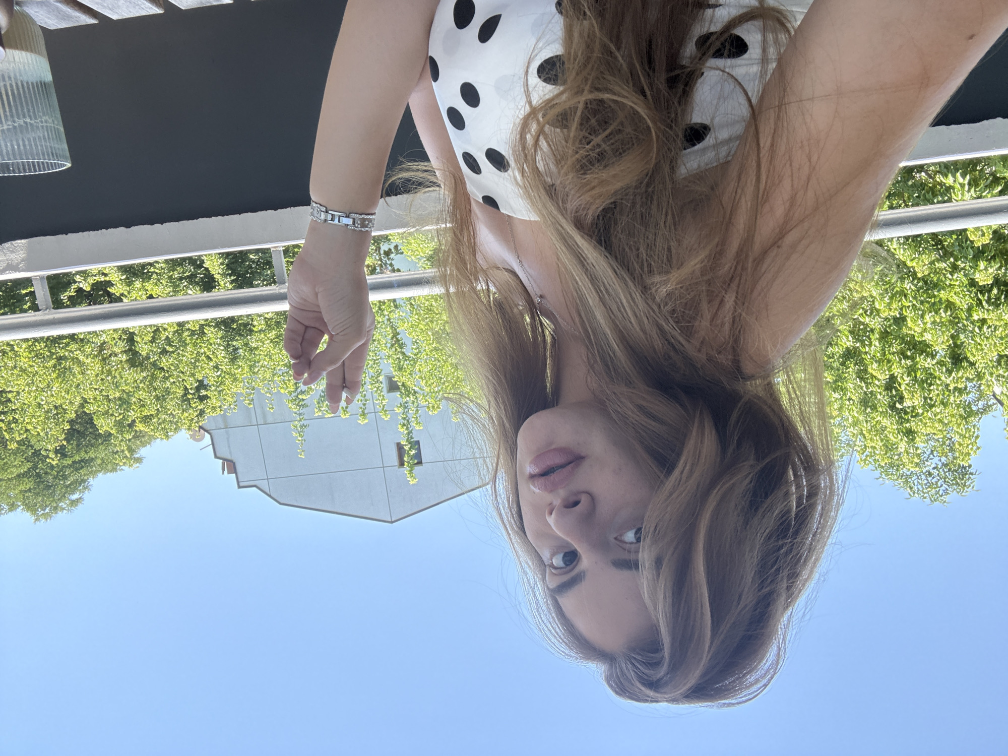 A young woman with long, wavy blonde hair is taking a selfie outdoors. She is wearing a white strapless top with black polka dots, a silver necklace, and a silver bracelet on her right wrist. Her right hand is raised, with her index finger and thumb forming a circle. She is looking at the camera with full lips and dark eyes. Behind her, there is a clear blue sky, green trees, a modern white building with a red-tiled roof, and a balcony railing. The environment suggests a sunny day in an urban setting. The image does not provide any specific details to determine the location in Plovdiv, Bulgaria.
