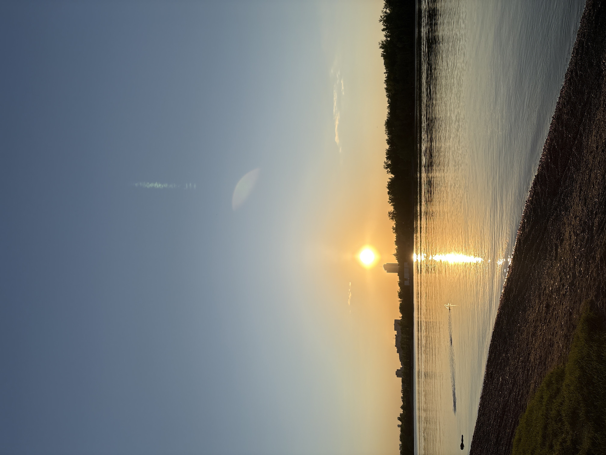 The image depicts a serene sunset over a wide body of water, likely a river or large lake, with an urban landscape visible on the distant bank. The sun, a bright white orb with an intense golden glow, is positioned low in the sky on the right side of the frame, casting a shimmering reflection across the water surface towards the viewer.
On the far bank, a line of dense green trees forms a dark silhouette against the bright sky and water, interspersed with several taller buildings or skyscrapers. The water itself appears calm, with gentle ripples reflecting the golden light of the setting sun.
In the foreground, a pebbled or sandy shore slopes gently into the water, with patches of green grass visible on the closest edge. A small, white, swan-shaped boat or pedal boat is floating on the water in the middle distance, directly in the path of the sun's reflection, creating a distinct silhouette. Closer to the foreground shore, a dark, indistinct silhouette of what might be a person or another object is visible in the water. The sky transitions from a deep blue on the left to a warm, clear golden-orange hue near the sun. A subtle lens flare is visible in the upper left portion of the sky.
The scene captures the peaceful atmosphere of a sunset. No visible text is present in the image. Given the provided context, the location is Krasnoyarsk, Russia, suggesting this is a view of the Yenisei River.