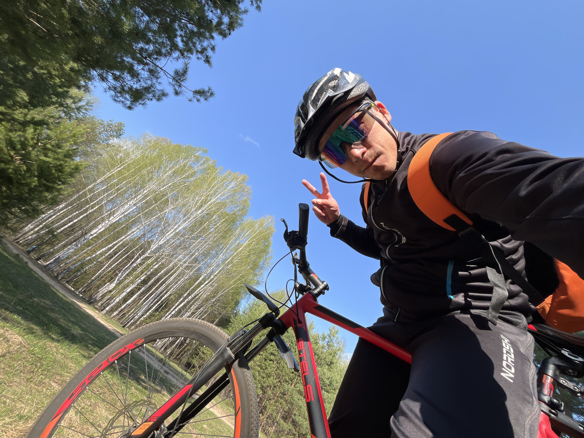 The image shows a man on a red and black mountain bike in a wooded area. He is wearing a black helmet and mirrored sunglasses, as well as a black long-sleeved jacket and dark pants. An orange backpack is visible over his right shoulder. He is holding up a peace sign with his left hand and appears to be taking a selfie. The background includes a dirt path and a forest with many birch trees. The sky is blue. The location is likely in a wooded area near Chaikovsky, Russia, based on the visible landscape features and the angle of the shot.