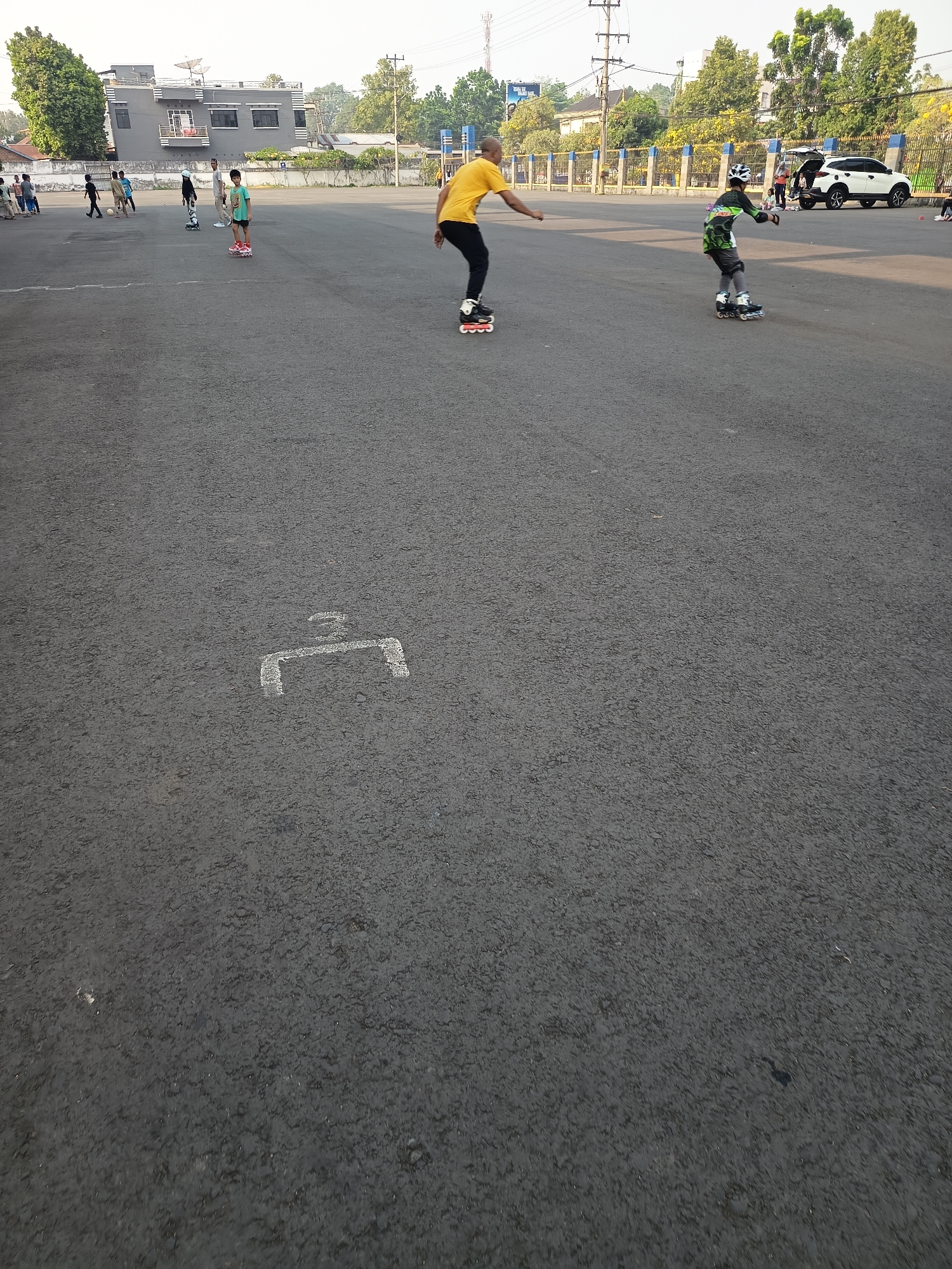 The image captures an outdoor scene in a paved area, possibly a parking lot or a sports court, with a building and trees visible in the background. In the foreground, a white painted marking on the asphalt surface resembles a stylized '7' or a bracket. Further into the scene, two individuals are rollerblading. One, wearing a yellow shirt and black pants, is in motion, with rollerblades on his feet. Another, dressed in a green and black patterned long-sleeved shirt, gray shorts, and protective gear including a helmet and knee pads, is also rollerblading. Several other people and a white SUV are present in the middle ground. A fence runs along the edge of the paved area, separating it from a strip of greenery and more distant buildings and trees. The sky is clear and overcast, suggesting daytime. The presence of a white SUV with its trunk open and people gathered around it might indicate a recreational or community event. The building in the background features a modern design with satellite dishes on its roof. The overall impression is a public space being utilized for leisure activities. While the image itself does not explicitly state the location, the context provided about Lubuklinggau, Indonesia, suggests this scene is taking place in a city within that region.