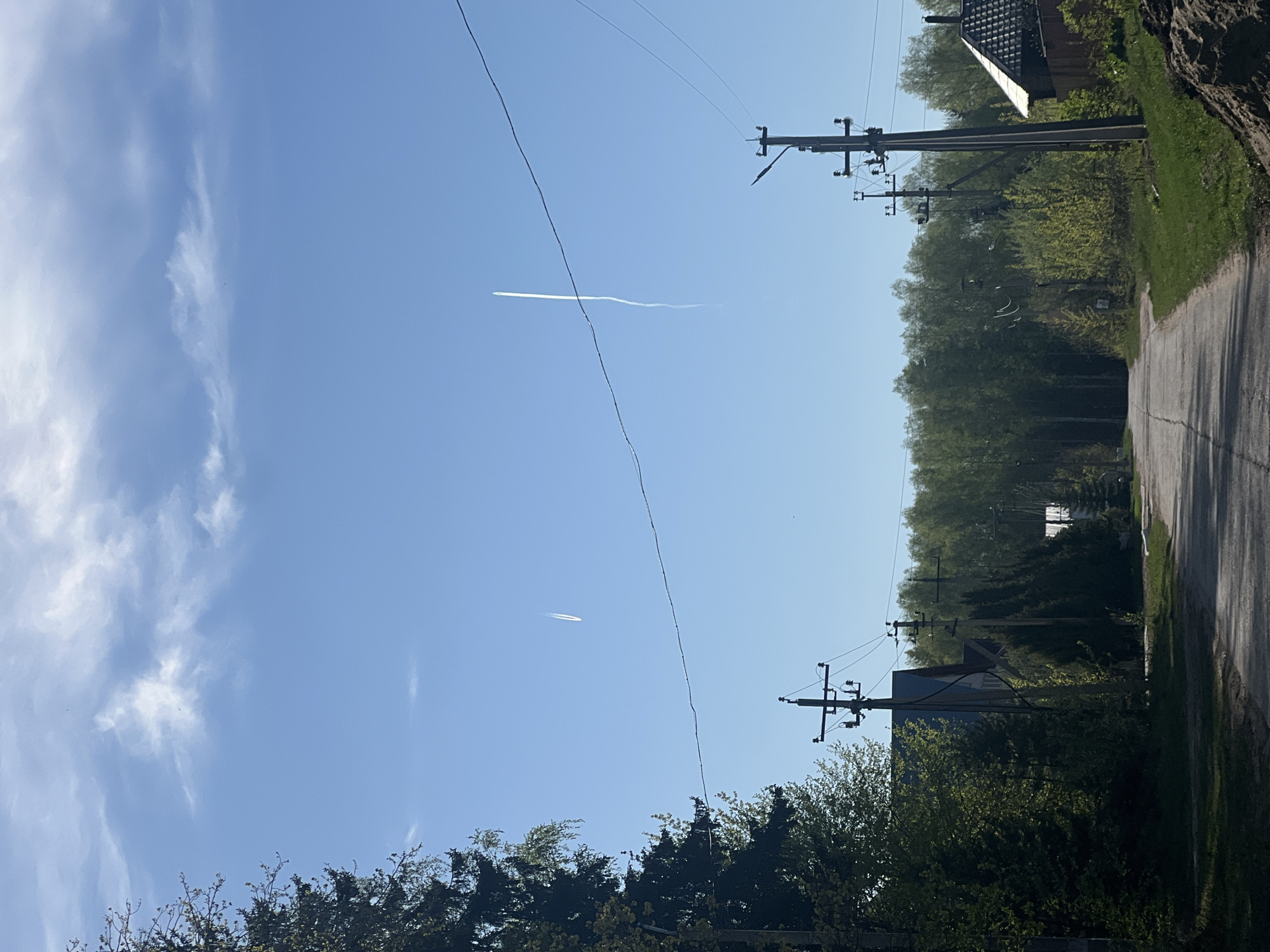 The image depicts a rural scene on a clear day in Noviy, Russia. A paved road, lined with trees and utility poles, stretches into the distance. A residence is partially visible on either side of the road. The sky is blue with light cloud formations. Two contrails, potentially from aircraft, are visible. Power lines run across the sky.