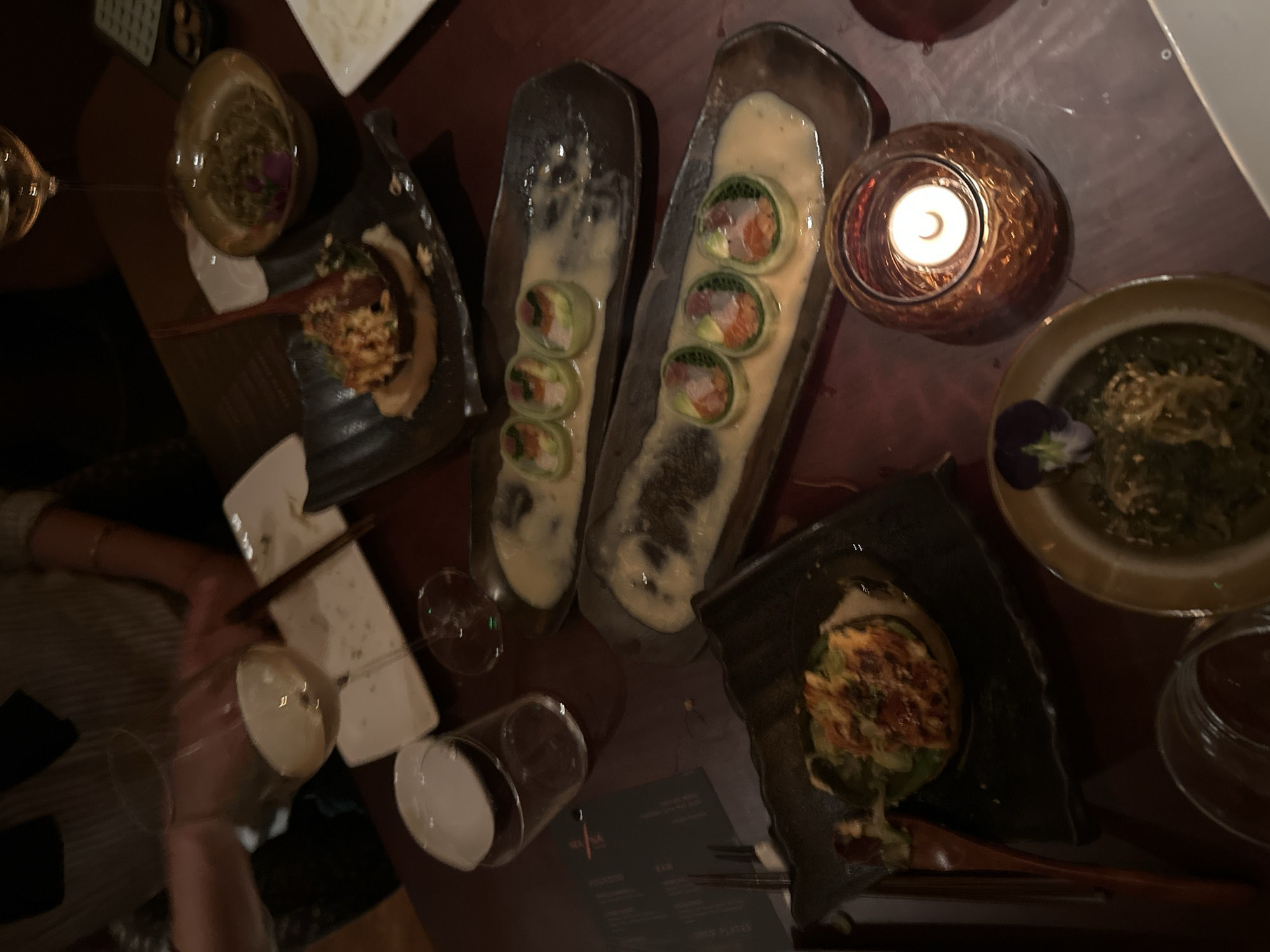 Captured from an overhead angle, the image showcases a dimly lit dining table laden with an assortment of dishes, indicative of a meal shared by multiple individuals. Predominantly, the cuisine appears to be Asian-inspired, with sushi rolls artfully arranged in oblong dishes, interspersed with creamy sauces. Another dish features a mound of food, possibly avocado with a topping, served on a dark, uniquely shaped plate, while bowls contain dishes resembling seaweed salad and other Asian delicacies. The presence of wine glasses, some filled and some partially emptied, suggest the consumption of beverages. A candle, encased in a patterned glass holder, casts a warm glow across the scene. The environment seems to be a restaurant, potentially in the city of London, United Kingdom, given the style of cuisine and the setting. Parts of human arms and hands are visible, implying people are in attendance, though their actions are not directly discernible from the image.