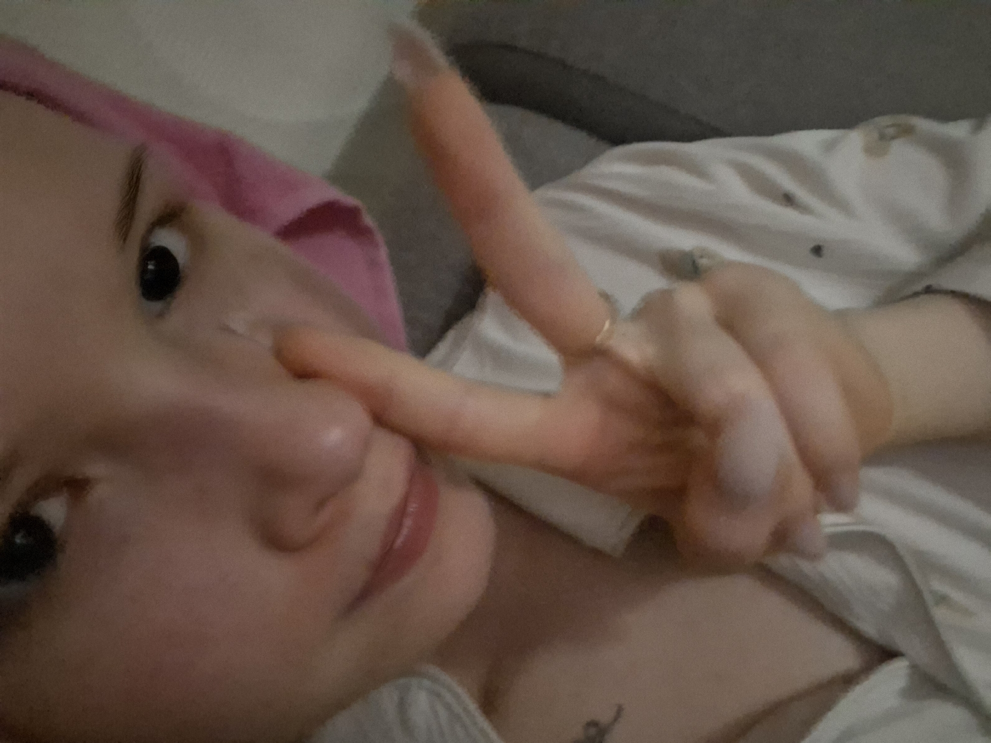 A person, likely female, is shown in a close-up, high-angle selfie, seemingly lying down or reclining in an indoor setting in Chaikovsky, Russia. She has fair skin, dark eyes, and pink-tinted lips, looking directly towards the camera. A pink towel or similar cloth is wrapped around her head, and she wears a light-colored, possibly white or cream-colored, top or pajamas with subtle patterns.
Her right hand is prominently featured, making a "peace" sign (V-sign) with her index and middle fingers. The index finger is playfully placed against her nose. A simple ring is visible on her ring finger. A small, dark tattoo, appearing to be an intricate design or script, is visible on her chest below her neck. The background is indistinct, showing a gray surface, possibly a couch or bed headboard, consistent with a relaxed, casual home environment. The lighting is soft and ambient, suggesting an indoor setting without direct sunlight. The image has a slightly grainy texture. No visible text is present.