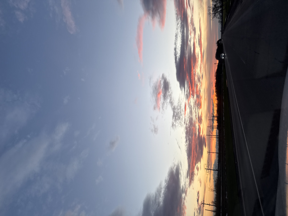 A sunset sky filled with a variety of clouds tinged with orange and pink hues stretches across a landscape. Utility poles line the horizon, creating a sense of depth. A road occupies the right side of the image, with two cars visible in the distance. The perspective suggests the photo was taken from inside a vehicle. The sky transitions from a pale blue to vibrant shades of orange and red closer to the horizon.