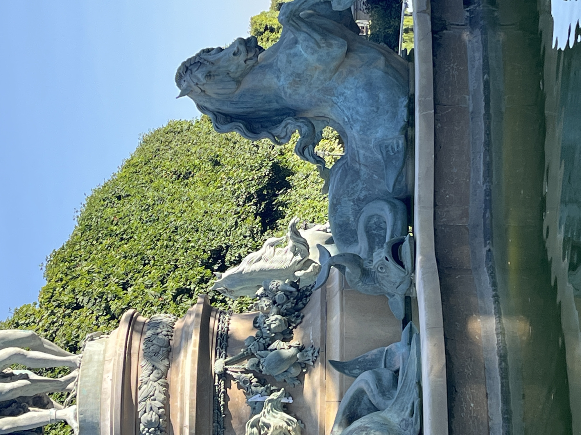 This image depicts a bronze sculpture of a mythical sea horse, a hippocampus, emerging from a fountain in Paris, France. The sculpture is detailed, with the horse’s mane flowing and its musculature defined. It is positioned in a fountain surrounded by lush green foliage. The water in the fountain is green and reflects the sky. The sculpture is situated at the base of a larger monument, parts of which are visible at the top left of the frame, featuring classical human figures. The scene is bathed in natural sunlight, suggesting it was taken during the day.