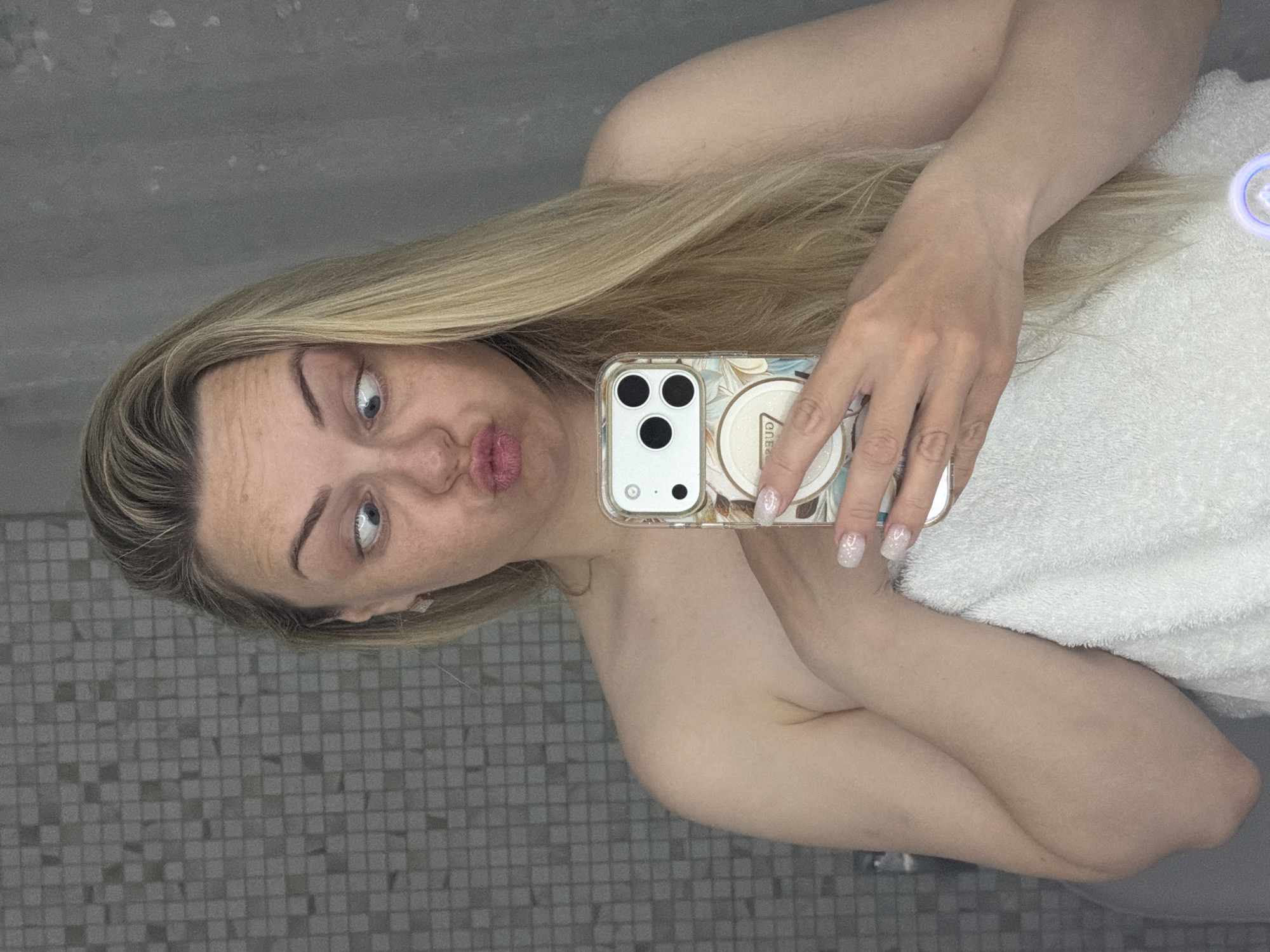 A close-up, sideways mirror selfie captures a woman with blonde hair and fair skin. She is looking upwards and slightly to the side with wide blue eyes, making a "kissing" or "pouty" face towards her smartphone. Her hair is long and falls over her shoulders and arm. She appears to be wearing a white towel, draped over her chest and shoulder, suggesting she might be in a bathroom or after a shower.
She holds a smartphone with a clear, patterned case (featuring floral or abstract designs) and a circular pop socket or stand on the back. The phone's triple camera system is visible. Her fingernails are manicured with a glittery, light-colored polish.
The background, visible in the mirror's reflection, consists of a grey tiled wall with small, square tiles, suggesting an indoor setting like a bathroom. The overall impression is one of a playful or casual moment captured in a self-portrait. Visible text on the circular pop socket is partially obscured and reversed, but appears to be "ONE".