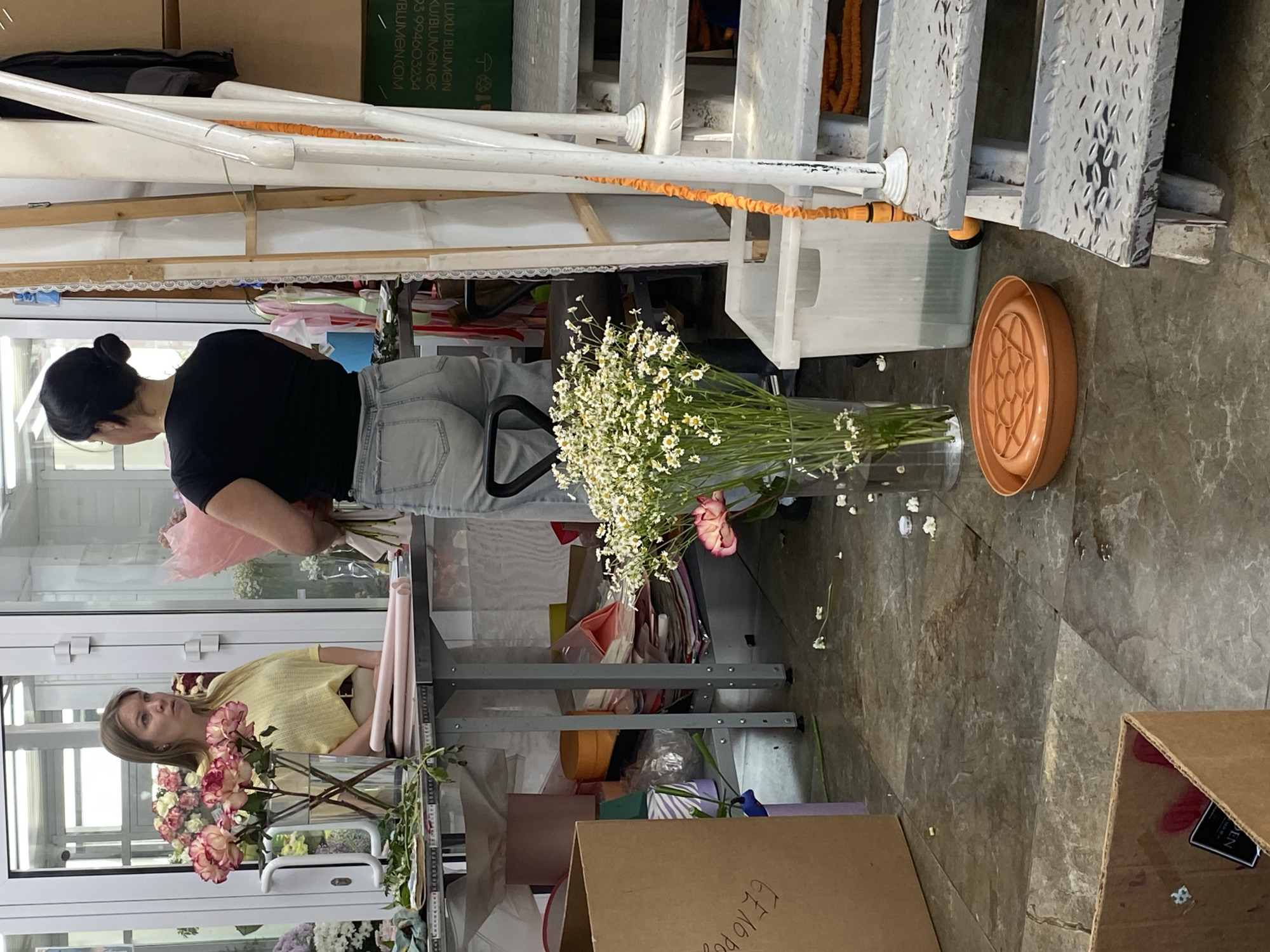 This appears to be an indoor setting, possibly a florist shop or a workspace dealing with flowers, located in what is suspected to be in Yekaterinburg, Russia. Two women are visible; one wearing a black t-shirt and jeans is seen from the back, handling flowers. The second woman is wearing a yellow top. There are vases of flowers in front of the woman with the yellow shirt. The scene includes bouquets and arrangements of flowers, a grey metal workbench, a cardboard box, and scattered floral debris on the floor. There are also several white stairs. The lighting appears bright, suggesting the presence of natural light sources or studio lighting.