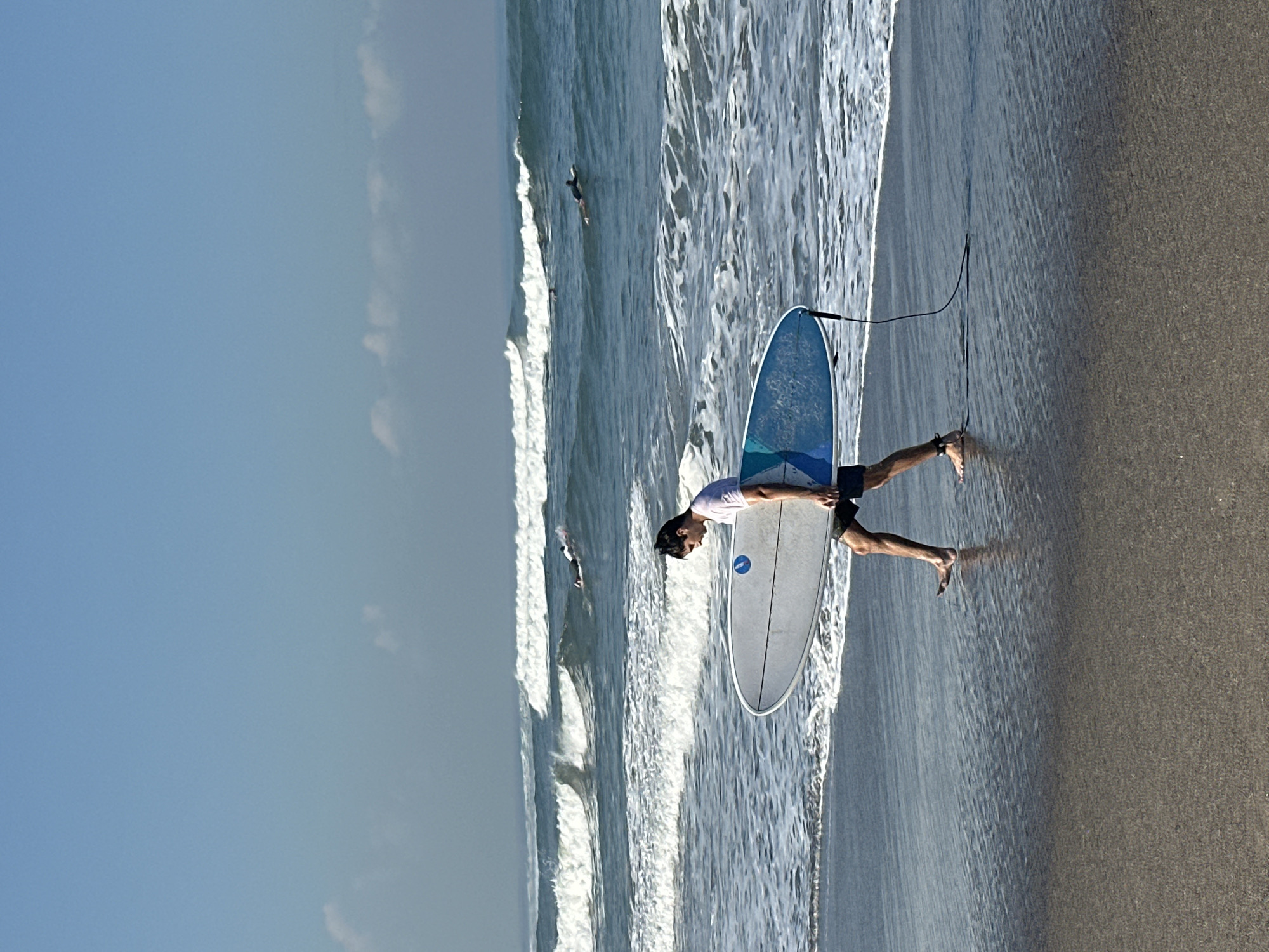 A man in a white t-shirt and dark shorts walks on a wet sand beach in North Kuta, Indonesia, carrying a white surfboard with a blue and white design. Waves crash on the shore as other surfers are visible in the ocean in the background under a clear blue sky with a few wispy clouds. The water reflects the sky and waves, creating a shimmering effect on the wet sand.
