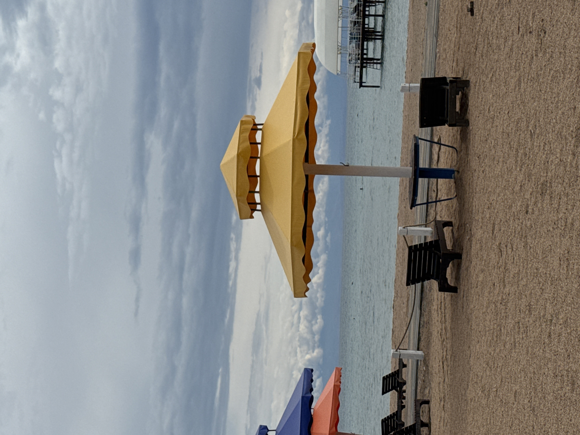 This image depicts a beach scene in Sary-Oy, Kyrgyzstan, on a cloudy day. The foreground is a sandy beach with several dark-colored lounge chairs. In the mid-ground, a large yellow umbrella stands over a blue table with a white pole. To the left, parts of blue and orange umbrellas are visible. The water in the background is calm and a pier with a covered structure extends into the water. The sky is heavily clouded with a gray and white pattern. There are no people clearly visible in the image, though a distant figure can be seen in the water.