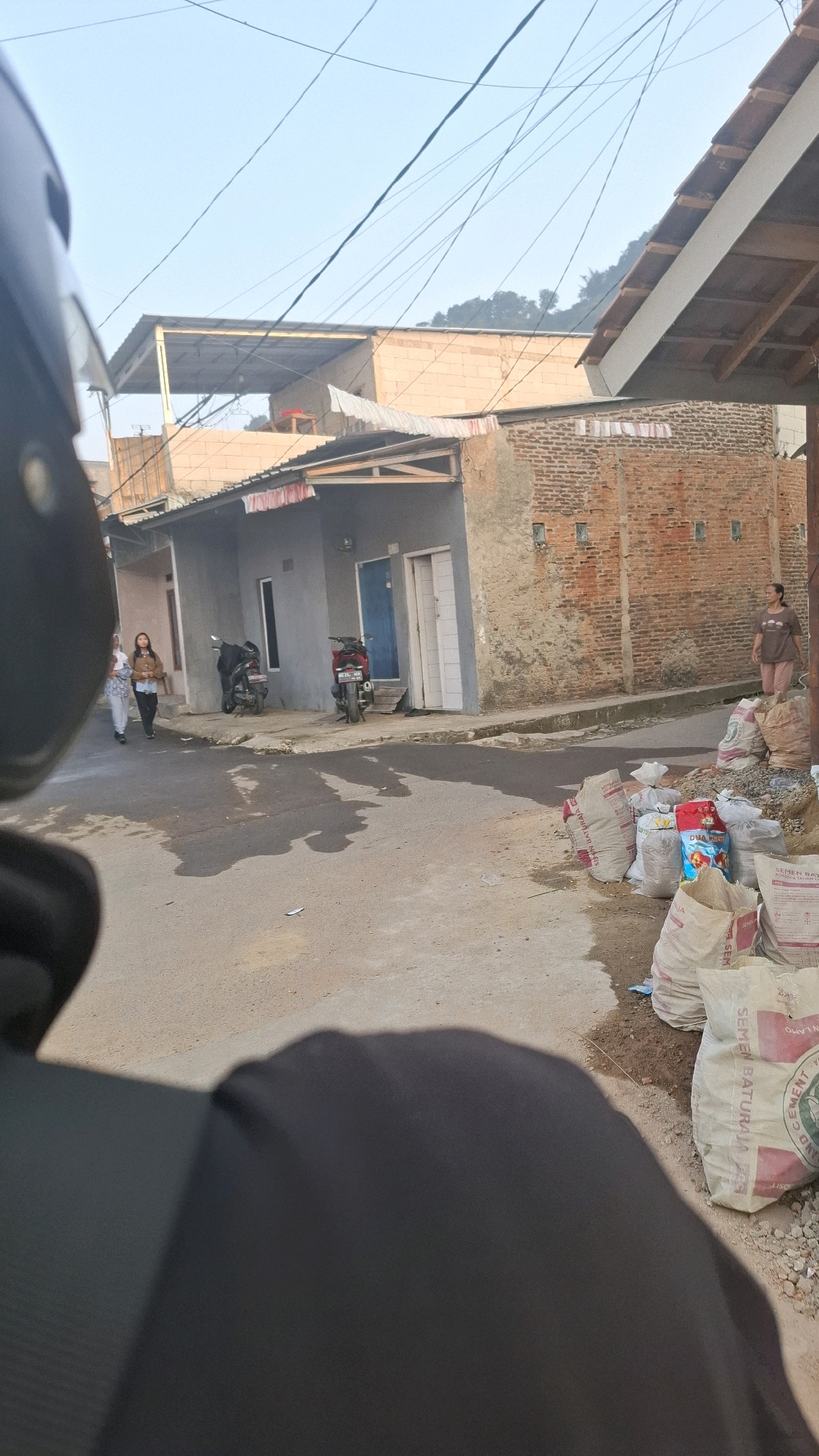 The image depicts a street scene in Bandar Lampung, Indonesia, on a clear day. In the foreground, the perspective suggests the viewer is seated on a motorcycle, with the rider's black helmet and shoulder area visible. The street is paved with asphalt and shows signs of wear and some puddles of water.
To the left of the street, two young women are walking. One is wearing a white top and striped trousers, and the other has on a brown jacket over a white top and dark trousers. Adjacent to them, parked on the side of the road, are two motorcycles.
Further down the street, on the left, is a small gray house with a blue door and two white doors. The house appears to have a corrugated metal roof. To the right of this house is a building constructed with orange bricks, showing signs of age and some plaster damage.
On the right side of the street, near the brick building, there is a pile of large sacks, some of which are white and branded with red and blue lettering, likely containing building materials such as cement. A woman in a brown t-shirt and pink trousers is standing near this pile, looking towards the left of the frame. The background shows more buildings, some with tiled roofs, and a hillside covered with trees. Overhead, several electrical wires are strung across the sky.