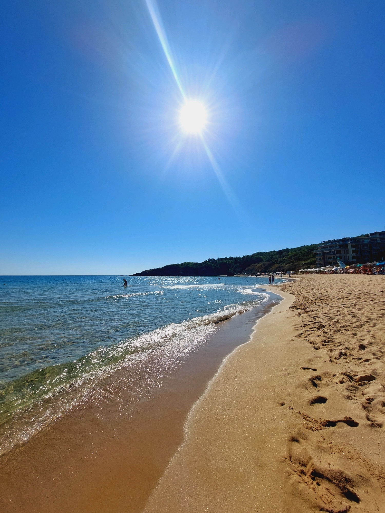 The image depicts a sun-drenched beach scene in Sozopol, Bulgaria. The foreground features golden sand with footprints, meeting the gentle surf of the clear, blue sea. The water is shallow and reflects the bright sunlight, creating a shimmering effect.
In the middle ground, a lone figure is visible standing in the water. Further down the beach, several people are walking along the shoreline and relaxing under umbrellas on the sand. A line of trees borders the back of the beach, and a multi-story building, likely a hotel or resort, is situated on a hill overlooking the coastline. The sky is a clear, vibrant blue, with the sun positioned high and casting a strong glare.