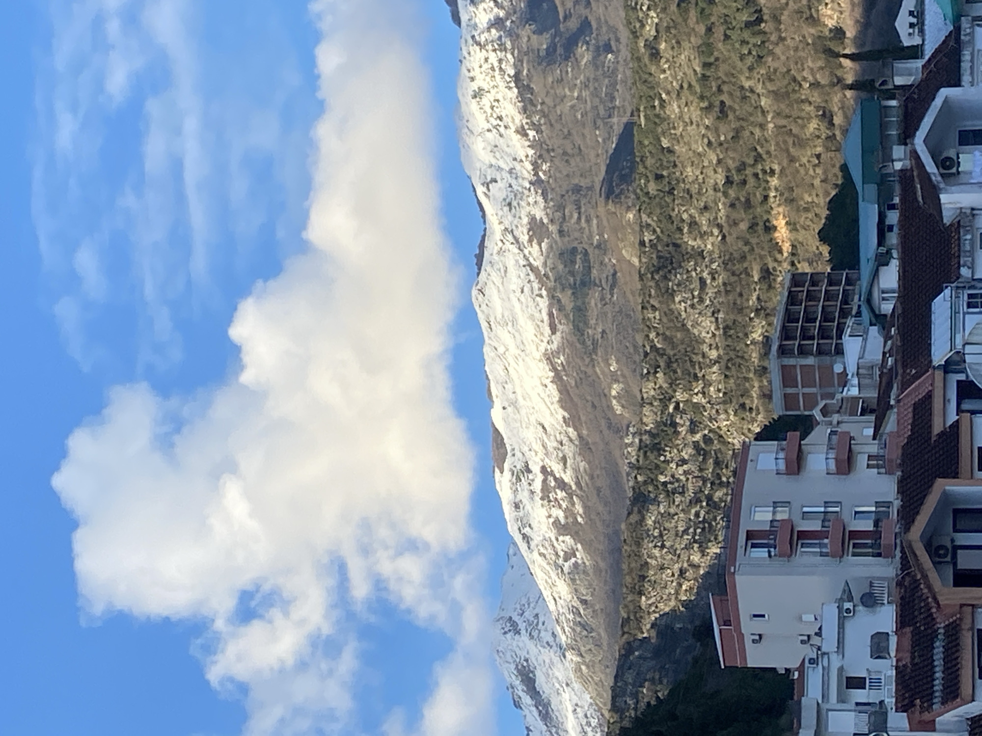 The image displays an elevated view of a residential area in Budva, Montenegro, backed by snow-capped mountains under a blue sky with scattered cumulus clouds.
In the foreground, a cluster of buildings comprises residential structures with characteristic reddish-brown tiled roofs and lighter-colored facades. A prominent five-story light-colored (beige/grey) apartment building with red-framed balconies and numerous windows is centrally located, exhibiting exterior air conditioning units. To its right, an unfinished multi-story building with exposed concrete framework is visible. Several satellite dishes are affixed to rooftops.
The midground transitions into lower hills and mountainous terrain, characterized by rugged, rocky surfaces and sparse, brownish vegetation, indicative of deciduous trees in a dormant state or sparse evergreen growth.
The background is dominated by a substantial mountain range with its peaks and upper slopes heavily covered in white snow. Below the snowline, the mountain slopes show areas of rocky exposure and scattered patches of vegetation.
The scene is well-lit by natural sunlight, suggesting daytime. No individuals, animals, or discernible activities are present.