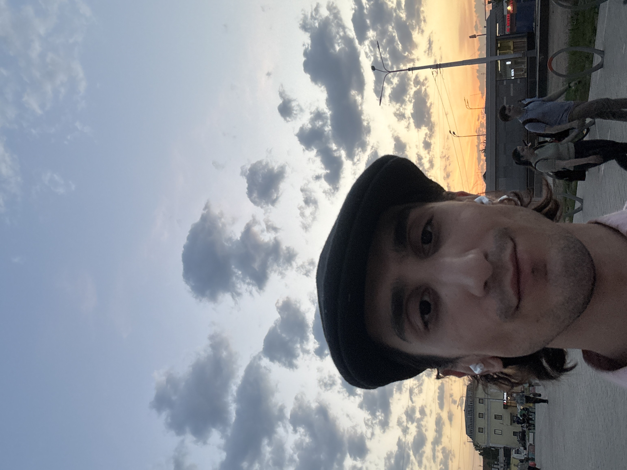 The image is a selfie of a man wearing a black hat, against a backdrop of a sky filled with clouds during a sunset. In the background, there is a street scene, including buildings, streetlights, and people walking. Based on the architectural style of the buildings, the location is likely in Moscow, Russia.
