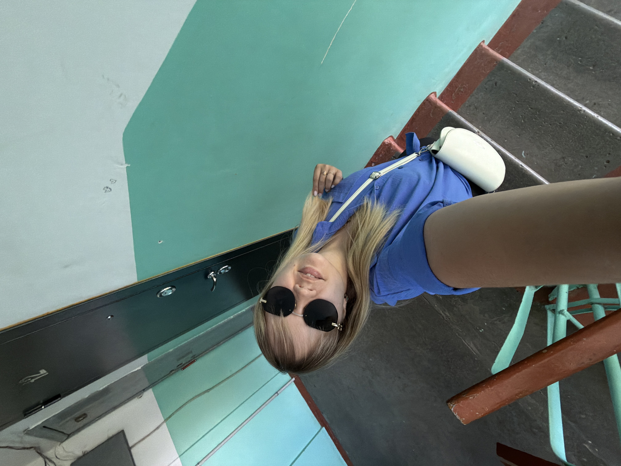 A young woman is taking a selfie on a staircase in what appears to be a residential building in Chaikovsky, Russia. She's wearing a blue shirt, sunglasses, and has a white bag. The staircase has painted metal railings in a faded blue-green color and the stairs themselves are concrete with some red trim. The walls of the building are painted in sections of white and a matching blue-green. A dark door or utility access panel is visible on the wall.