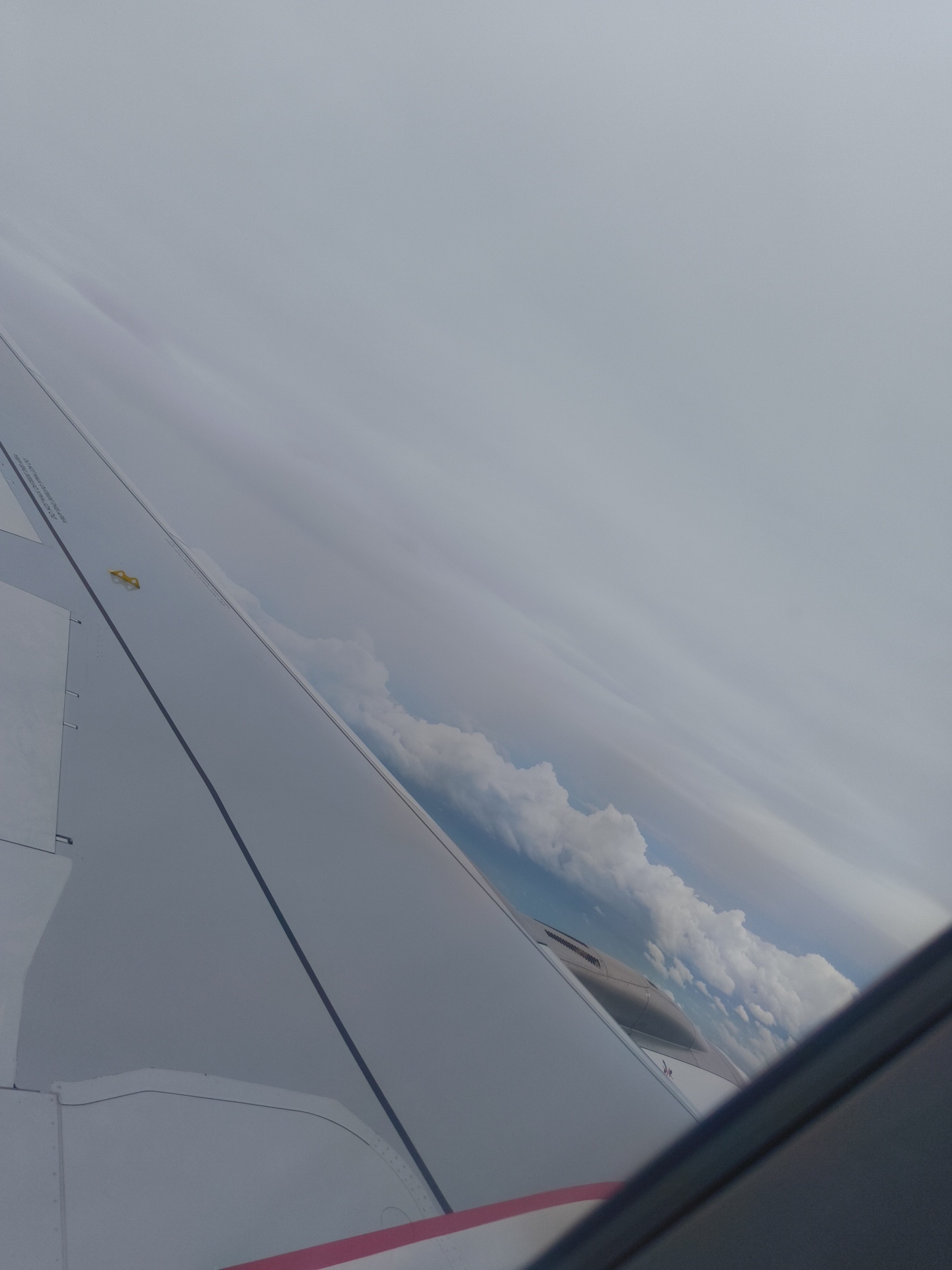 The image is taken from the window of an airplane, looking out over a wing. The wing is white with some gray lines and rivets visible. There is a small yellow object on the wing. Text is visible on the wing, but it is too blurry to read. In the distance, there are clouds and a blue sky. The time of day appears to be daytime, and the weather seems to be clear with some clouds. The setting is an airplane in flight. The image conveys a sense of travel and observation from above.