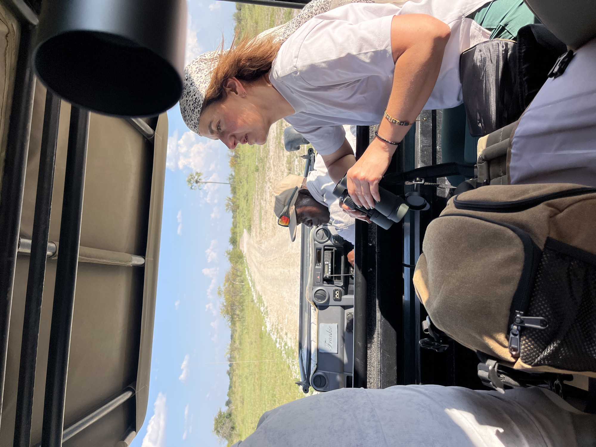 The image shows a woman with reddish-brown hair wearing a white t-shirt and patterned headscarf looking through binoculars from the back of an open-top safari vehicle. A Black man wearing a white shirt and a beige hat is visible in the driver's seat. The dashboard of the vehicle is visible, featuring a plaque reading "Frankie". The landscape outside the vehicle appears to be grassy plains, with a single palm tree visible in the distance under a blue sky with scattered clouds. The environment suggests a safari scene in a rural area, potentially in Gucta, Botswana. Backpacks are seen inside the car on the right. The woman and driver appear to be engaged in wildlife viewing.
