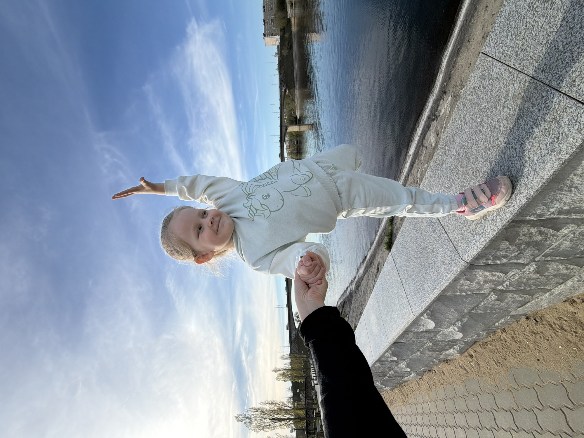 A photograph taken in Kamyshin, Russia, shows a young child, likely a girl, being held in a horizontal position as if flying. The child, dressed in a light-colored sweatsuit with a unicorn design, is suspended in the air, her body parallel to the waterline of a river visible in the background. Her right arm is extended outwards. She appears to be smiling at the camera. An adult’s arm and hand, wearing a dark sleeve, are visible at the bottom of the frame, holding the child’s hand for support. The background features a cityscape with buildings and a bridge spanning the river. The foreground includes a paved walkway. The sky above is blue with scattered clouds.