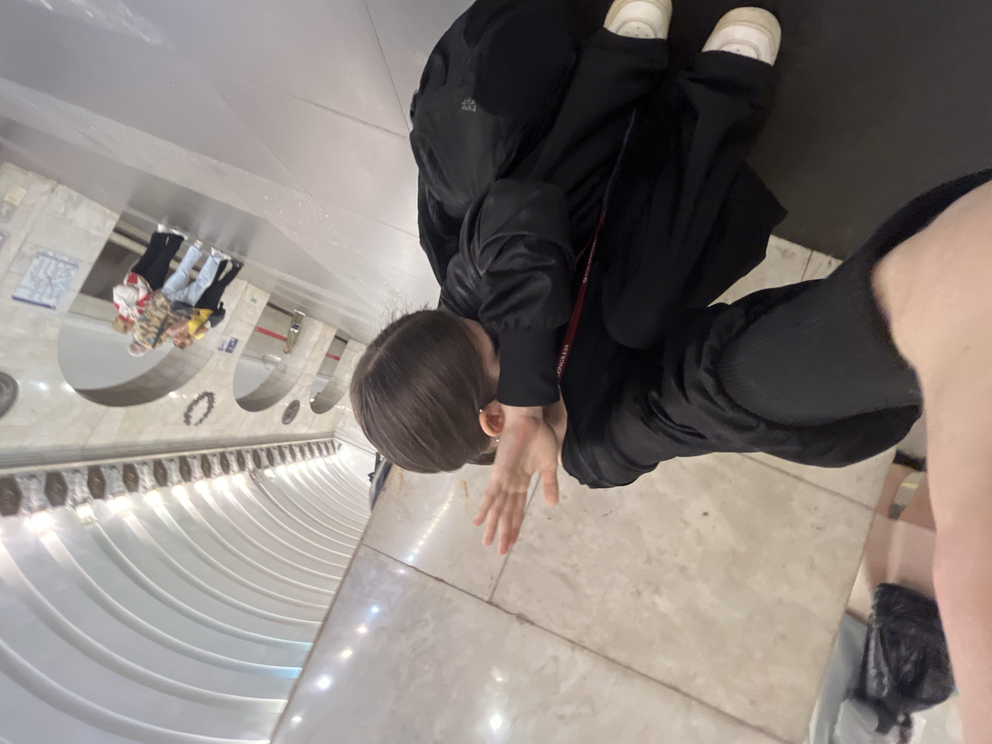 An overhead, slightly tilted shot captures a person, likely in an indoor public space, possibly a metro station in Saint Petersburg, Russia. The primary subject, with short brown hair, is dressed in black outerwear, black pants, and white sneakers, with a black backpack. They are positioned looking downwards, with one hand raised towards their face. A red lanyard or strap with the visible text "CONGRESS" hangs from their neck.
The floor beneath the subject is a light, reflective tiled surface. The background features distinctive architecture, including multiple curved white panels that suggest an escalator tunnel or a grand hall. Above these panels, arches and decorative elements are visible. Further in the background, a reflection on a polished surface reveals other people and what appears to be a map or signage on a wall, typical of a transport hub. The scene is illuminated by artificial lights, casting reflections on the shiny surfaces. The unique architectural style is consistent with the ornate and distinctive metro stations found in Saint Petersburg.