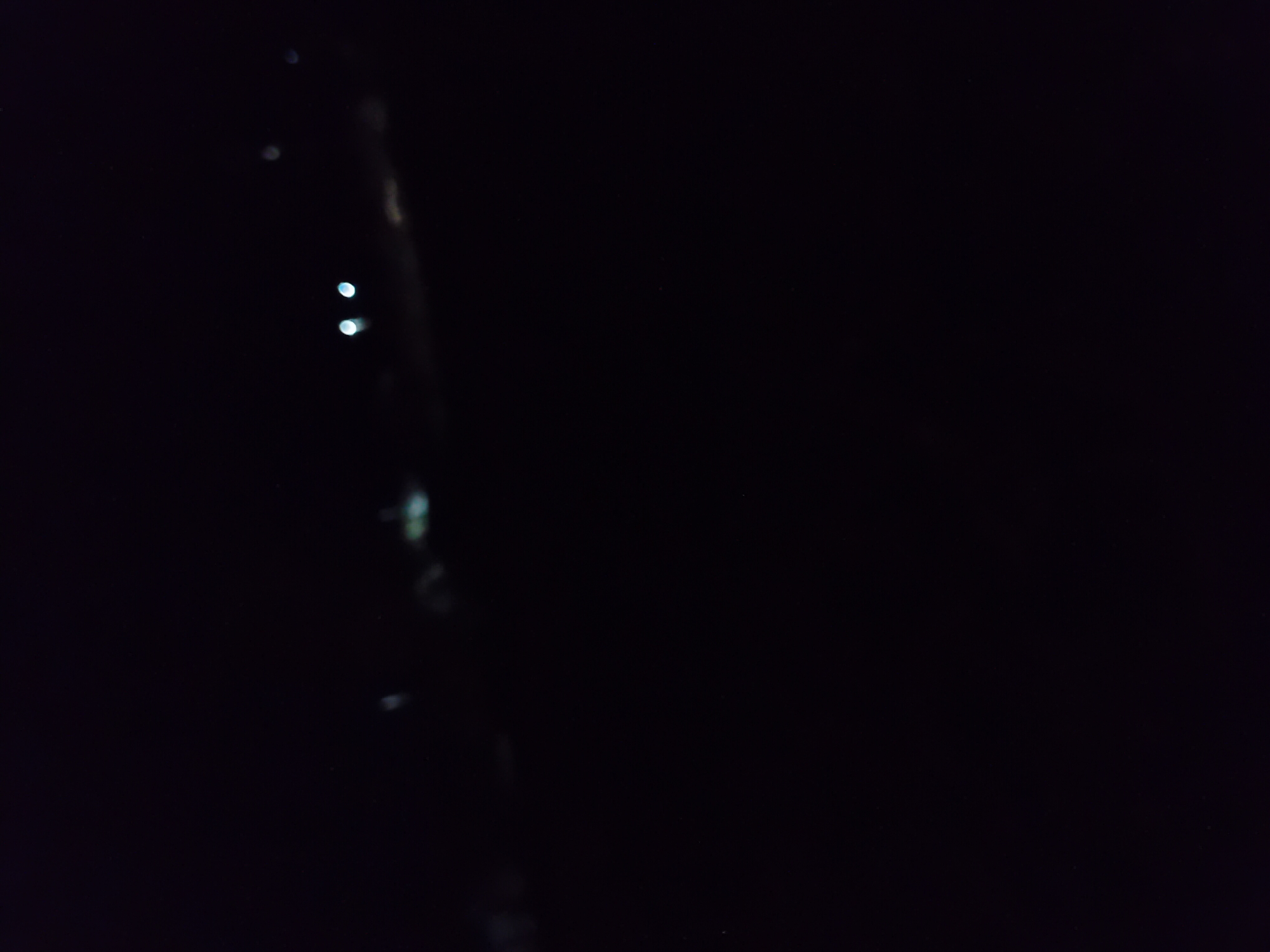 The image is a dark, blurry photograph taken at night, likely from a slightly elevated position. In the background, several out-of-focus lights are visible, appearing as bright, circular smudges. Two prominent, bright white lights are clustered together in the center-left of the frame, suggesting they might be headlights of a vehicle. Other fainter lights are scattered across the upper portion of the image, potentially indicating distant streetlights or windows. The overall scene is dark and indistinct, with the majority of the image being black. There is no discernible information about people, specific objects, actions, or interactions. The environment is not identifiable beyond being an outdoor, nighttime setting with artificial lights. No specific location within Jauro Yinu, Nigeria can be determined from the visual information provided.