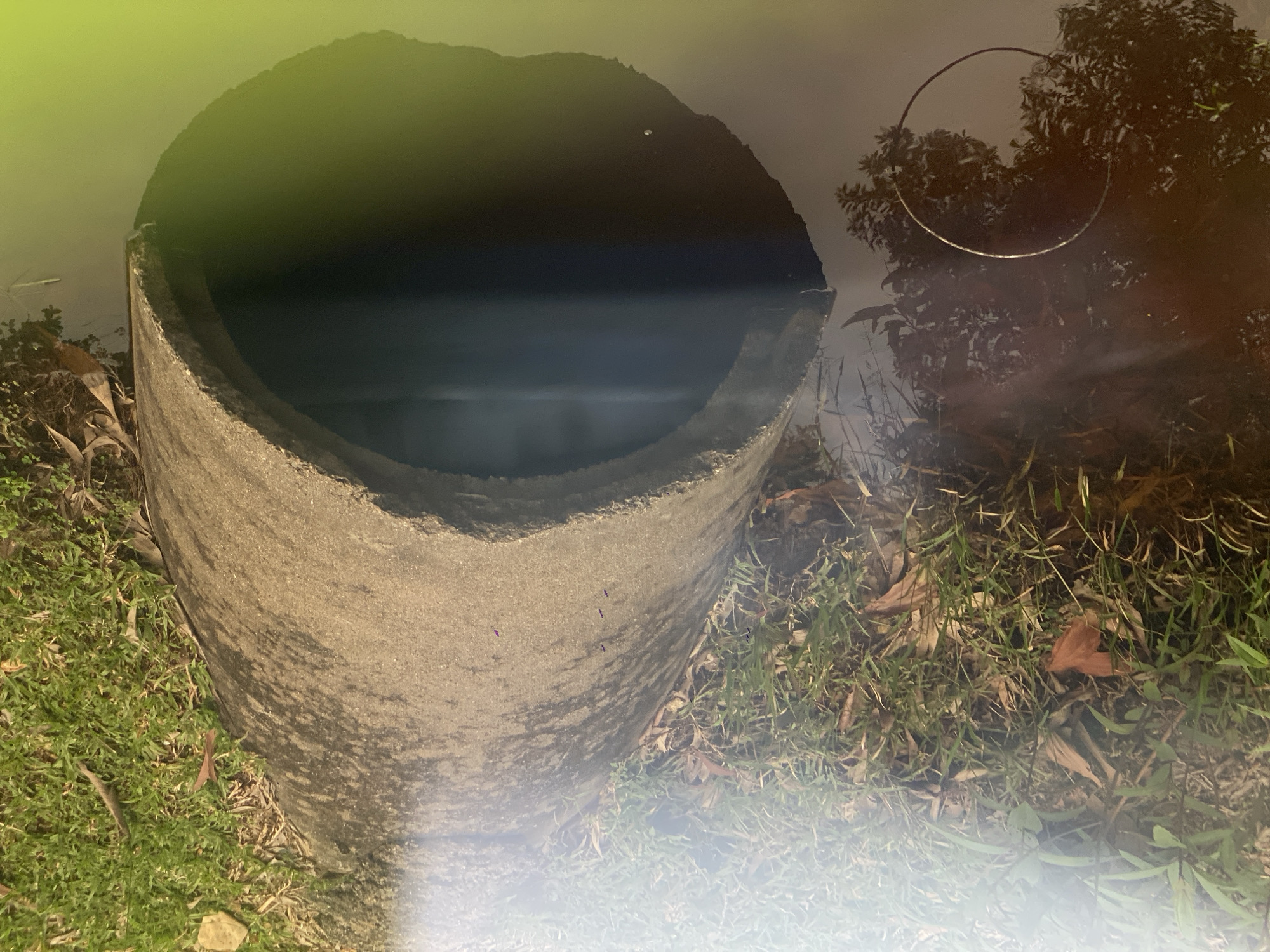A concrete pipe with an arched opening is partially submerged in dark, still water. The opening of the pipe leads into a shadowy interior. The surrounding environment consists of green grass and dry leaves scattered on the ground. The water reflects the surrounding vegetation, creating a blurred image on its surface. A faint circular object is visible in the water to the left of the pipe. The scene appears to be outdoors, possibly near a drainage area or waterway within Ko Siboya, Thailand.