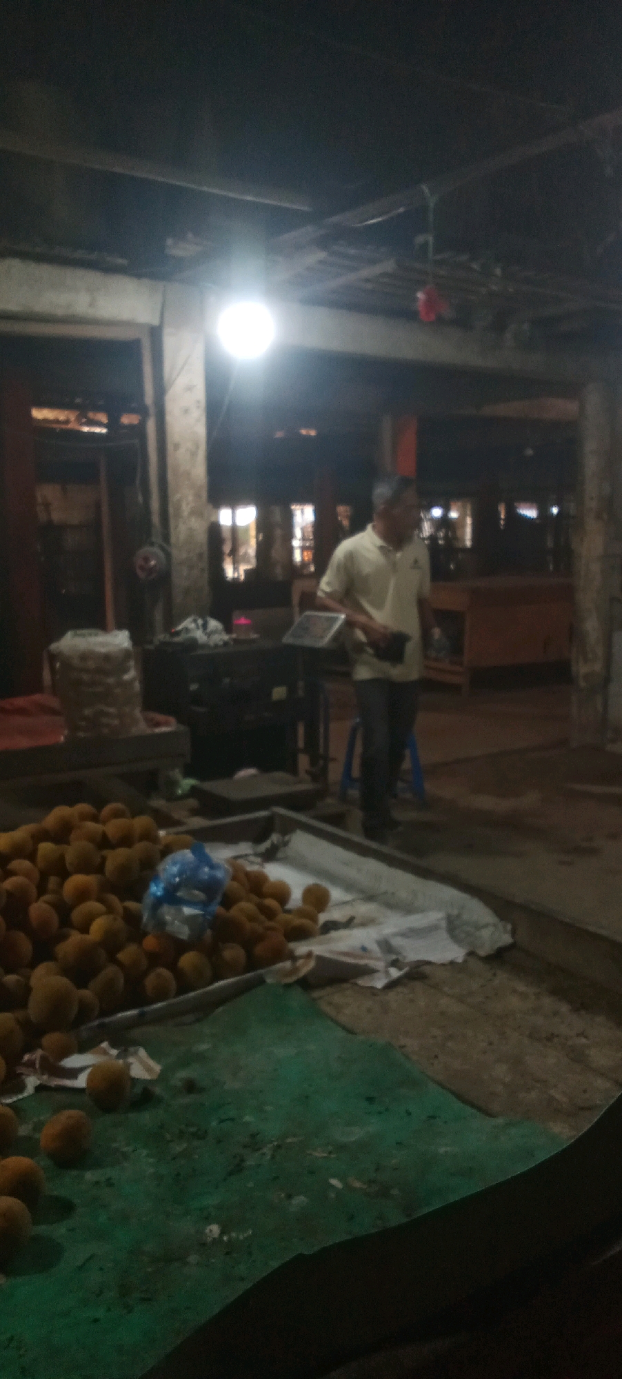 A man in a light-colored polo shirt and dark pants stands near a fruit stand in what appears to be an open-air market at night. The market is dimly lit by a single bright overhead light. In the foreground, a large pile of round, orange fruits, possibly loquats, is displayed in a metal bin. A blue plastic bag sits on top of the fruit. To the left, there is another pile of a white, fibrous material, possibly straw or packing material. Behind the man, there are dark wooden shelves and counters, suggesting additional market stalls. The ground is a mixture of dirt and concrete. The man is holding a dark object, possibly a wallet or phone, in his right hand. The overall scene depicts a typical night market setting in Karawang, Indonesia.