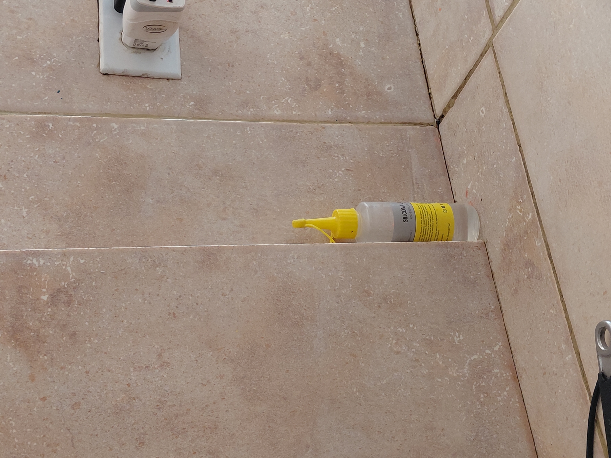 The image displays a close-up, static indoor scene, likely in Santa Ana, Venezuela, featuring various objects against a background of light brown, speckled ceramic tiles.
In the upper left, a white wall outlet is visible, into which a white and grey power adapter or charger is plugged. The brand "Drawey" is clearly legible on the adapter. Below this, the tiled wall forms a horizontal surface or shelf.
Resting on this tiled surface, in the center-right of the image, is a clear plastic bottle with a bright yellow cap and nozzle. The bottle contains a clear liquid. A yellow label wraps around the bottle, with the text "SILICONA LI" prominently visible. Below this, the words "USO GERAL" can be partially seen, indicating a general-purpose silicone liquid. Further small text and symbols are present on the label but are illegible due to resolution and angle.
The entire background consists of uniform light brown tiles with subtle darker speckles, creating a textured appearance. The grout lines between the tiles are visible, with some showing minor discoloration or dirt, particularly in the corner where two tiled walls meet on the right side of the image. On the far right edge of the image, a small metallic hook, possibly part of a hanger or fixture, is partially visible.
There is no activity depicted; the objects are stationary. The lighting suggests an indoor environment, possibly illuminated by artificial light or natural light from an unseen source.