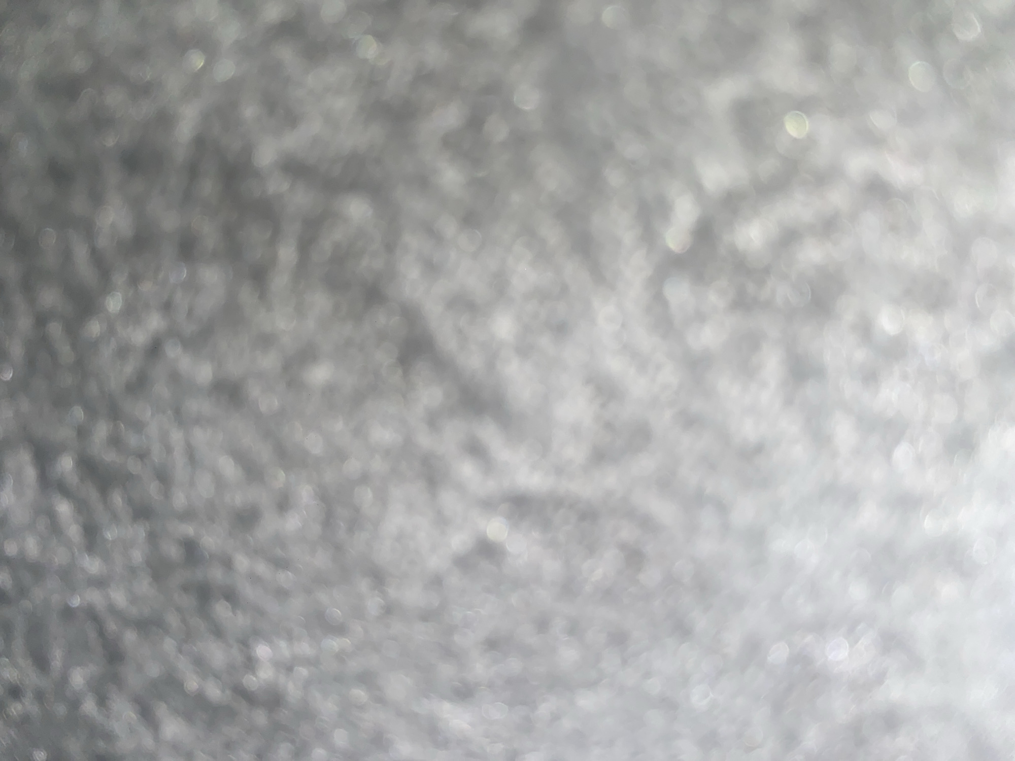 The image is an abstract, out-of-focus shot of a shimmering, textured surface. The dominant color palette consists of various shades of gray and white, with subtle hints of iridescent light reflecting off the surface. The texture appears granular and uneven, with small, circular points of light scattered throughout, suggesting a glittery or frosted material. There are no discernible objects, people, or actions present. The scene is devoid of any specific environmental context that would place it within Ablanitsa, Bulgaria, or any other location.