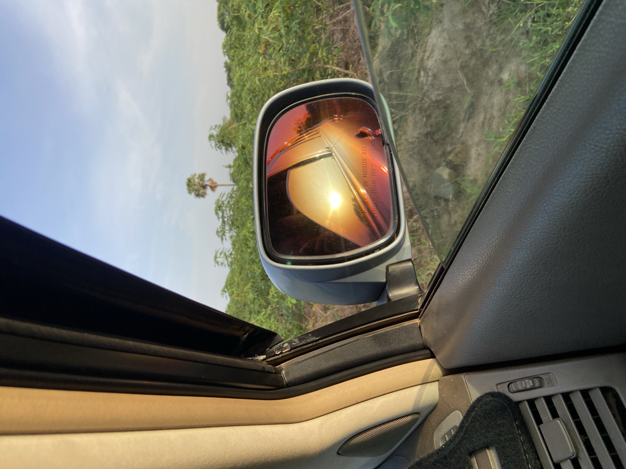 The image is taken from inside a car, focusing on the passenger-side mirror. The mirror reflects a golden sunset or sunrise with some vegetation. The background visible through the car window includes a dirt road and lush greenery, possibly agricultural land. In the distance, a single palm tree stands against a pale sky. The car's interior is visible, including the dashboard, air vents, and door panel. The scene suggests a location in a rural or semi-rural area near Mararaba, Nigeria. There are no people visible in the photo.