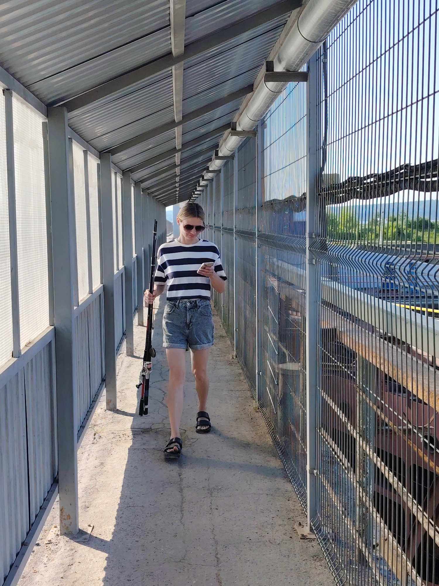 A woman walks along an enclosed pedestrian bridge in Chaikovsky, Russia. She holds a fishing rod in her left hand and a phone in her right. The bridge has a corrugated metal roof and is enclosed by metal fencing on both sides. Outside the fence on the right, a train and landscape can be seen.