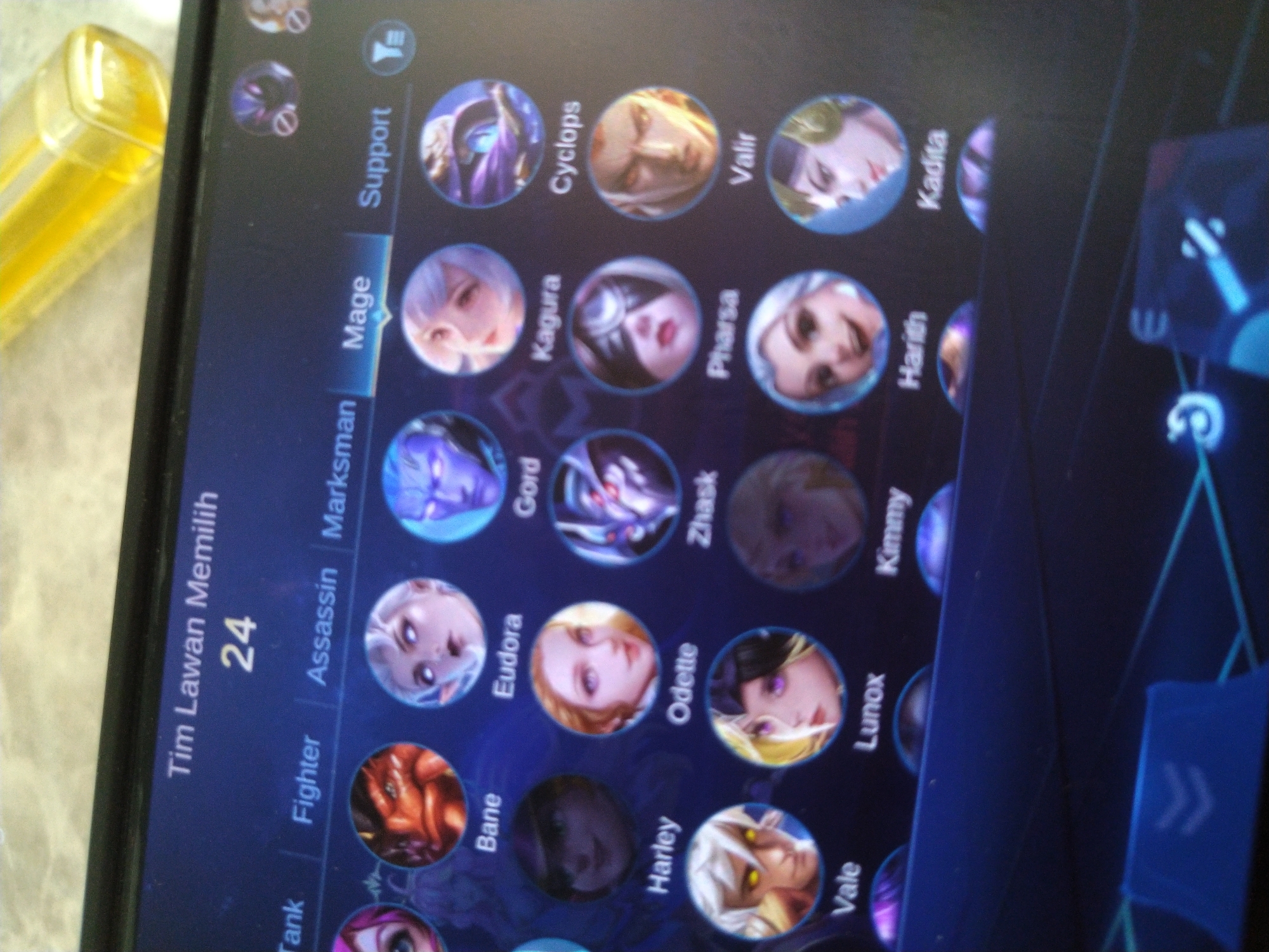 The image shows a close-up of a tablet screen displaying a character selection interface from a mobile game, likely "Mobile Legends: Bang Bang". The screen is oriented horizontally and the interface is in Indonesian. At the top, it reads "Tim Lawan Memilih" (Opponent Team Picks) with the number "24" below it, possibly indicating the number of available characters or a player count. Below this header, different hero role categories are listed: "Tank", "Fighter", "Assassin", "Marksman", "Mage", and "Support". A selection of hero portraits with their names underneath is visible. Notable heroes displayed include Bane, Eudora, Gord, Kagura, Cyclops, Harley, Odette, Zhask, Pharsa, Valir, Vale, Lunox, Kimmy, Harith, and Kadita. The bottom of the screen shows part of the game's UI with blue accents and icons. The tablet itself is positioned on a textured surface, possibly a table, and a portion of a yellow object, likely a lighter, is visible in the upper right corner of the frame. The image is taken at a slight angle, suggesting it was captured by a person holding a camera or another device. There is no visual information in the image that would allow for the identification of the location in Matangkruet, Indonesia.
