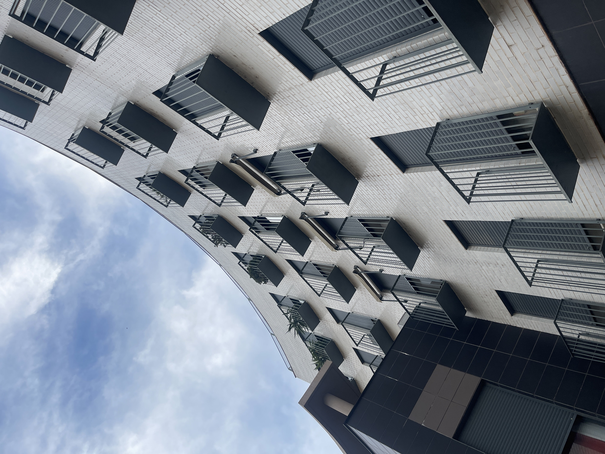 The image displays the modern, curved facade of a multi-story building in Manises, Spain, captured from a low-angle perspective looking upwards. The building's exterior is primarily clad in light-colored, off-white bricks. Numerous small, cantilevered balconies with dark grey metal railings and platforms project uniformly from the facade. Each balcony is situated in front of a window fitted with dark grey, closed roller shutters. Several balconies on the upper-left portion of the structure contain green potted plants or foliage. A distinct architectural section on the lower-left features a contrasting dark grey tiled facade. An additional prominent architectural element on the upper-left includes a dark, flat overhanging structure supported by a visible column. The sky above is partially overcast with scattered clouds and visible patches of blue, containing a minute dark speck, possibly a bird, in the upper-center. The scene is lit by diffused, natural light, indicating an overcast day.
