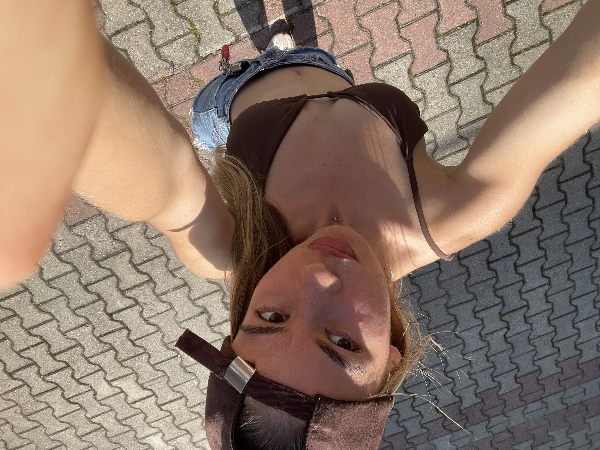 A young woman is captured in a high-angle, downward-facing selfie on a sunny day. She has light-toned skin, blonde hair, and dark eyes, looking directly upwards at the camera with a neutral to slightly intense expression. She is wearing a dark brown baseball cap backward, a dark brown bra-style top with thin straps, and light blue distressed denim shorts that are unzipped or loosely worn, exposing her lower abdomen and a small scar above her navel. A thin silver-colored necklace with a small pendant is visible. A keychain with keys and a red fob hangs from her right hip pocket. She is standing on interlocking concrete pavers, which are predominantly grey with interspersed sections of faded reddish-brown, indicating a paved outdoor area. Strong sunlight casts distinct shadows on the ground and highlights her features.