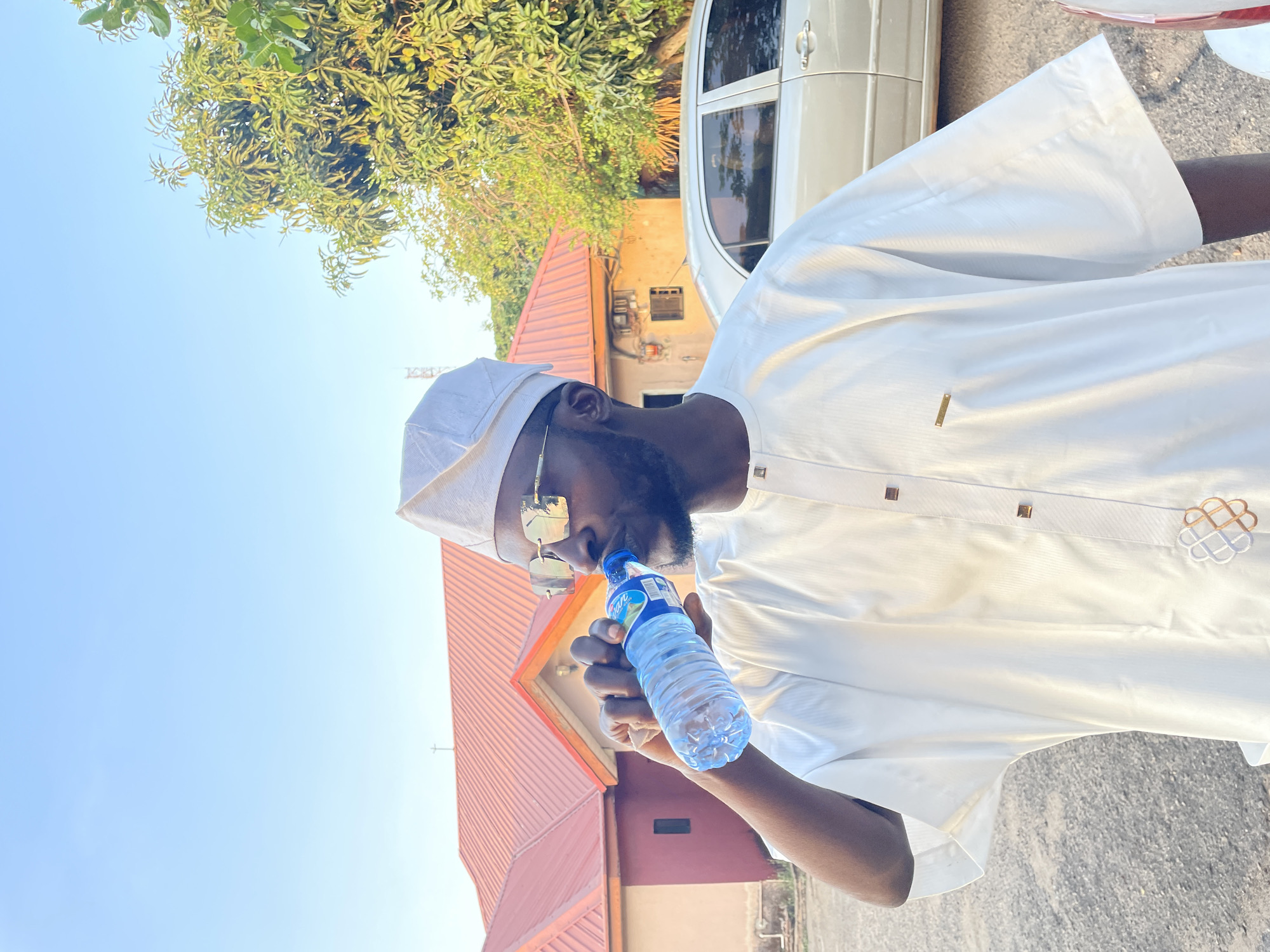 A man is drinking bottled water outside. He is wearing a white traditional outfit and a matching white cap. He also has sunglasses on. There's a building with a red roof behind him, and a car is parked nearby. Trees are visible in the background, and the sky is blue. The location is assumed to be Jos, Nigeria, but there is no visual confirmation of the city.
