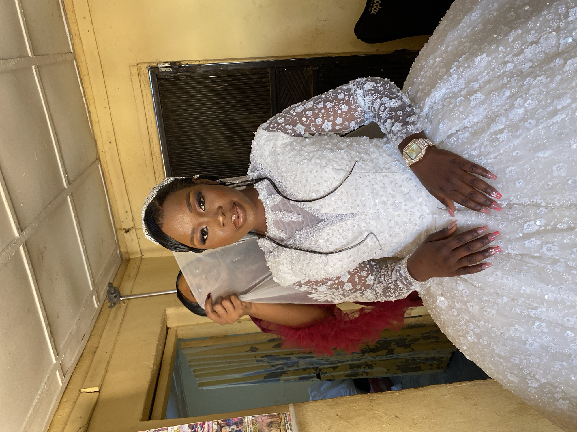 A woman in a white, long-sleeved, floral sequined wedding dress and bejeweled tiara poses for a picture indoors in Vom, Nigeria. She wears a gold watch on her left wrist, and her fingernails are painted pink. An unidentified person wearing a dark maroon feathered item holds a transparent white veil behind her. The background consists of a yellow wall, a dark door with horizontal slats, and various objects including an electrical box on the ceiling and a black bag. There is a window with yellow blinds, and various posters are attached to the wall.