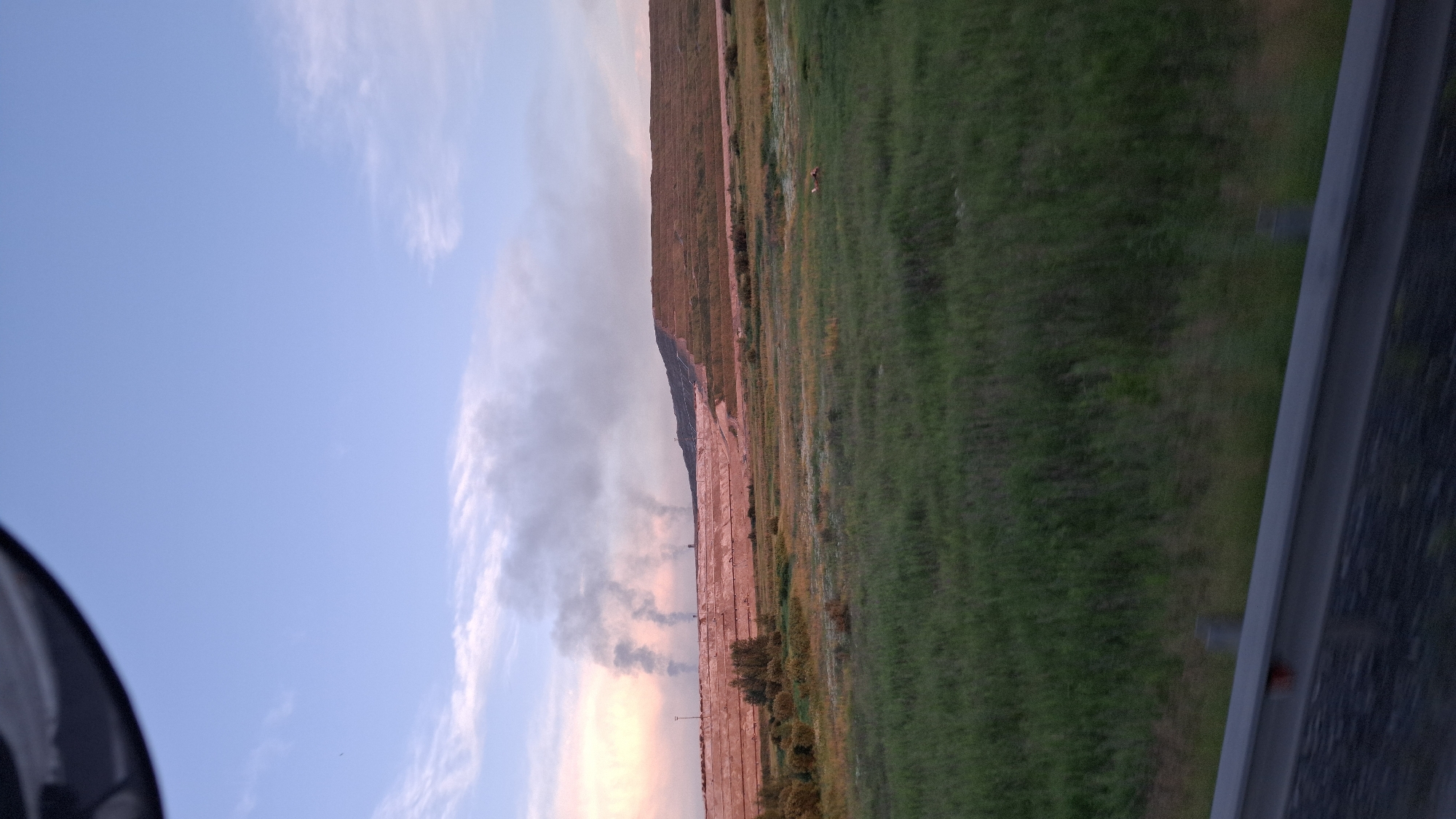 The image depicts a landscape scene with a clear sky, clouds, and a grassy field bordering a large embankment. The embankment appears to be part of a landfill or a construction site, with tiered layers and some vegetation growing on it. Smoke plumes are visible rising from the base of the embankment, suggesting an ongoing event such as a fire or industrial activity. In the distance, a few animals are present in the field. The sky displays a soft blend of blue and pink hues, indicating either sunrise or sunset. The foreground features a metallic railing, suggesting the viewer is observing the scene from an elevated platform or vehicle. There is no visible text in the image. The overall impression is one of a vast, somewhat desolate landscape experiencing an event that is generating smoke.
