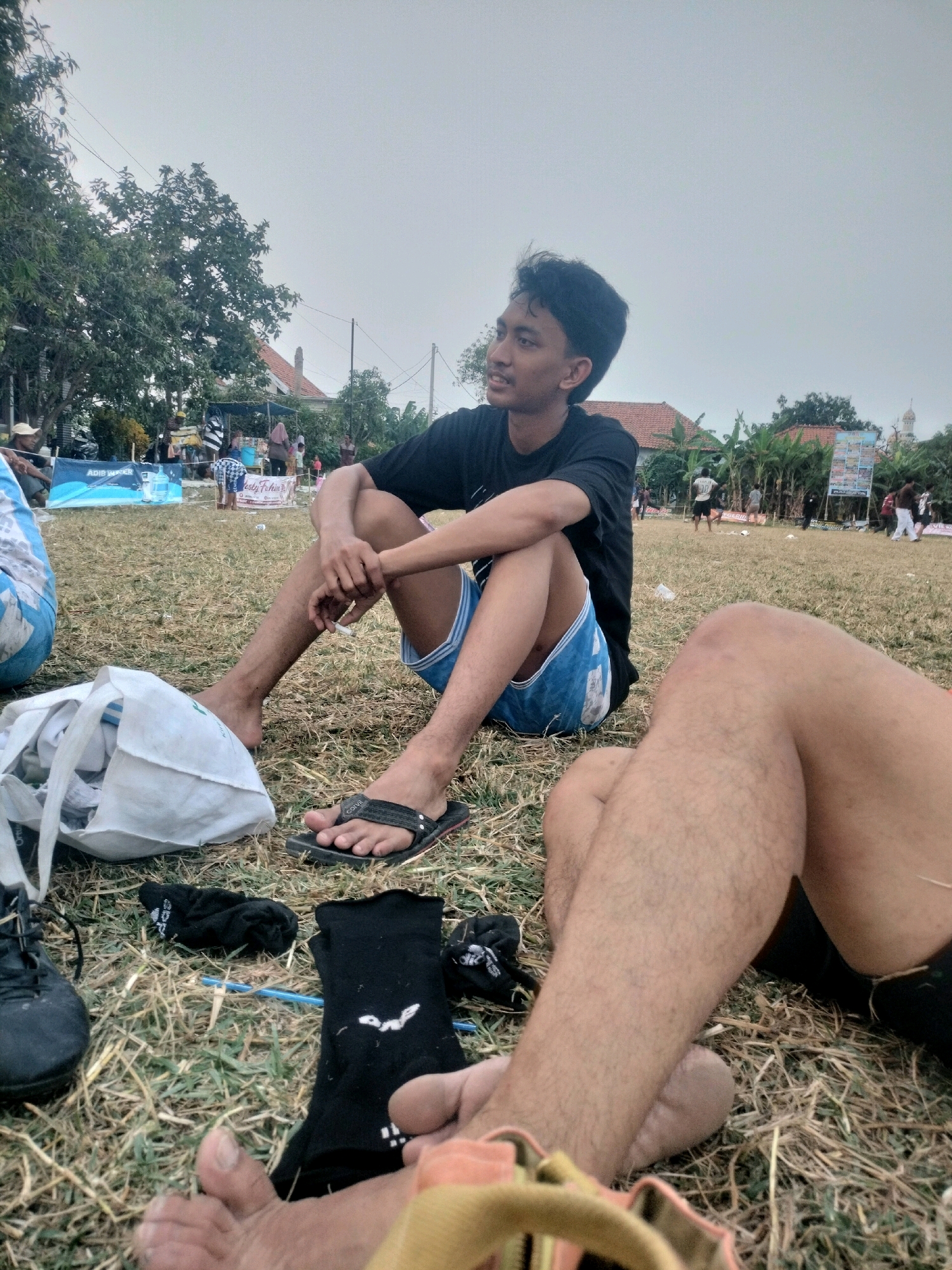 The image captures an outdoor scene with a group of people gathered in what appears to be a field or park. In the foreground, two individuals are seated on the dry grass. One person, a young male with dark hair, is wearing a black t-shirt and blue patterned shorts. He is sitting with his legs extended, and his hands are clasped. Adjacent to him, another person's legs are visible, also seated on the grass. Scattered on the ground are various items including a white bag, black socks, and a pair of black shoes.
In the background, there are several tents or stalls set up, suggesting a possible event or market. Various people are seen walking around and interacting in the distance. The environment is mostly flat with sparse vegetation and some trees in the mid-ground. The sky is overcast, indicating a cloudy day. The overall impression is a casual gathering in an outdoor setting.