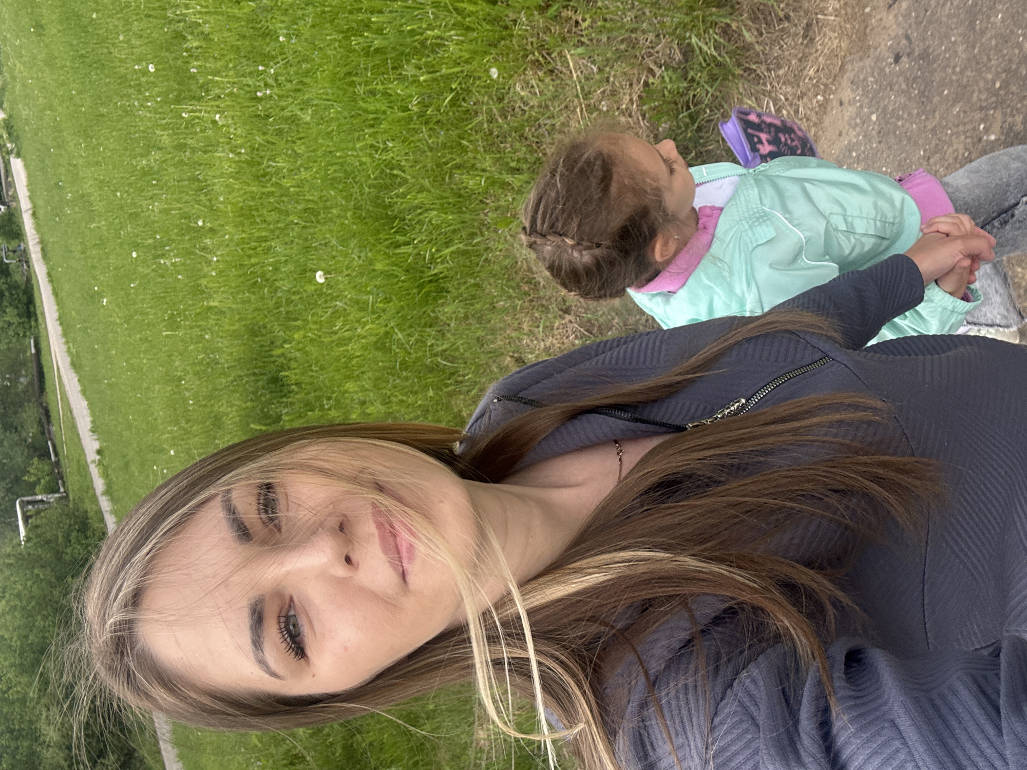 A woman and a child are on what appears to be a path next to a grassy field. The woman is wearing a grey zippered top, and her long brown hair is partially blonde. The child is wearing a green jacket with a pink lining and sitting next to the woman, with her hands interlocked. There is greenery in the background. It's difficult to confirm the location is Shevlyakovo, Russia without more contextual information.