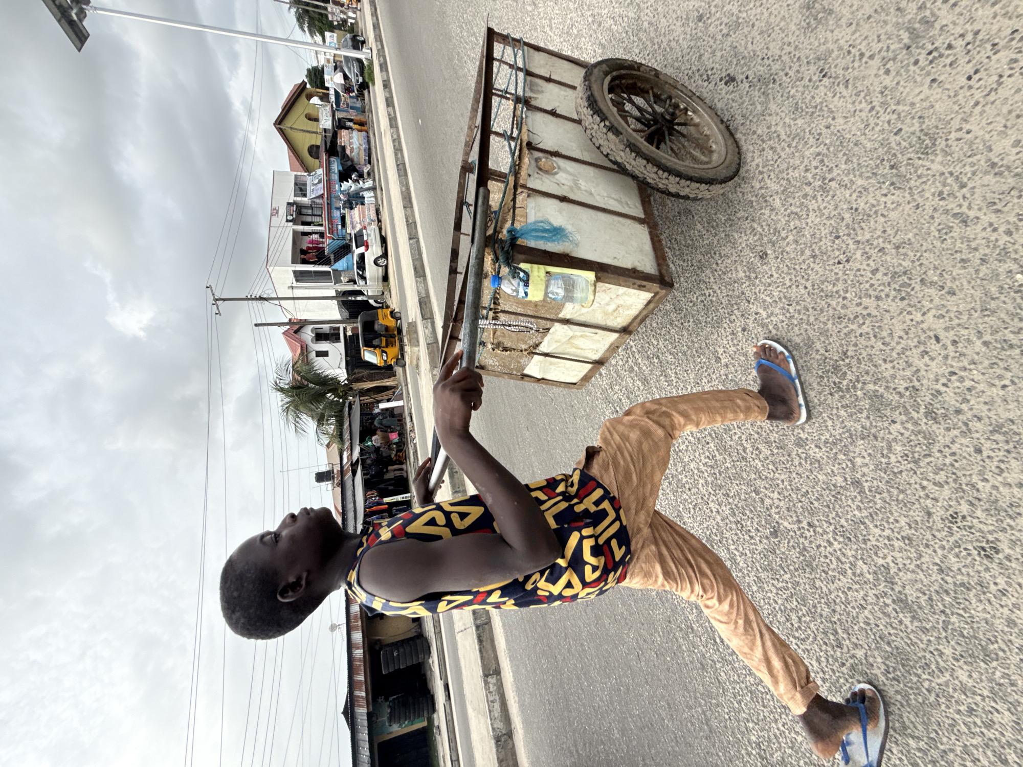 A young person is seen pulling a metal cart along a paved street in Ado, Nigeria. The cart has a spare tire attached and contains several water bottles. The person is wearing tan pants, a patterned shirt, and blue and white sandals. The road is flanked by buildings and shops, with various vehicles visible in the background. The sky is overcast.