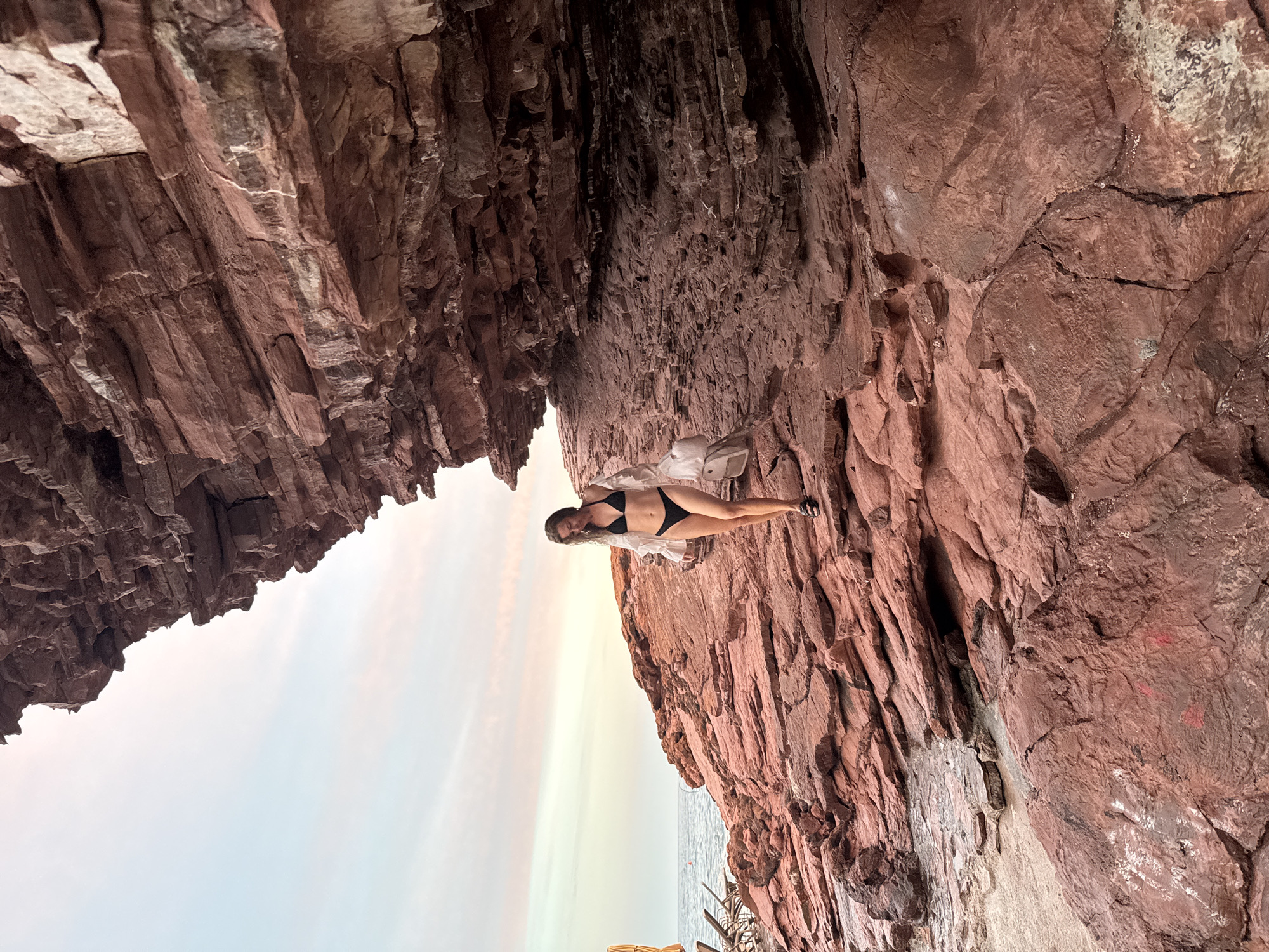 A woman wearing a black bikini and a white sheer cover-up walks on a rocky shoreline. The rocks are a reddish-brown color and appear layered, suggesting a geological formation. Behind the woman, the sea is visible with calm water under a pale sky with streaks of pink and orange. To the right of the woman, a large rock overhang creates a shaded area. The scene is located in Sveti Stefan, Montenegro, a coastal area known for its dramatic rock formations and scenic beaches.