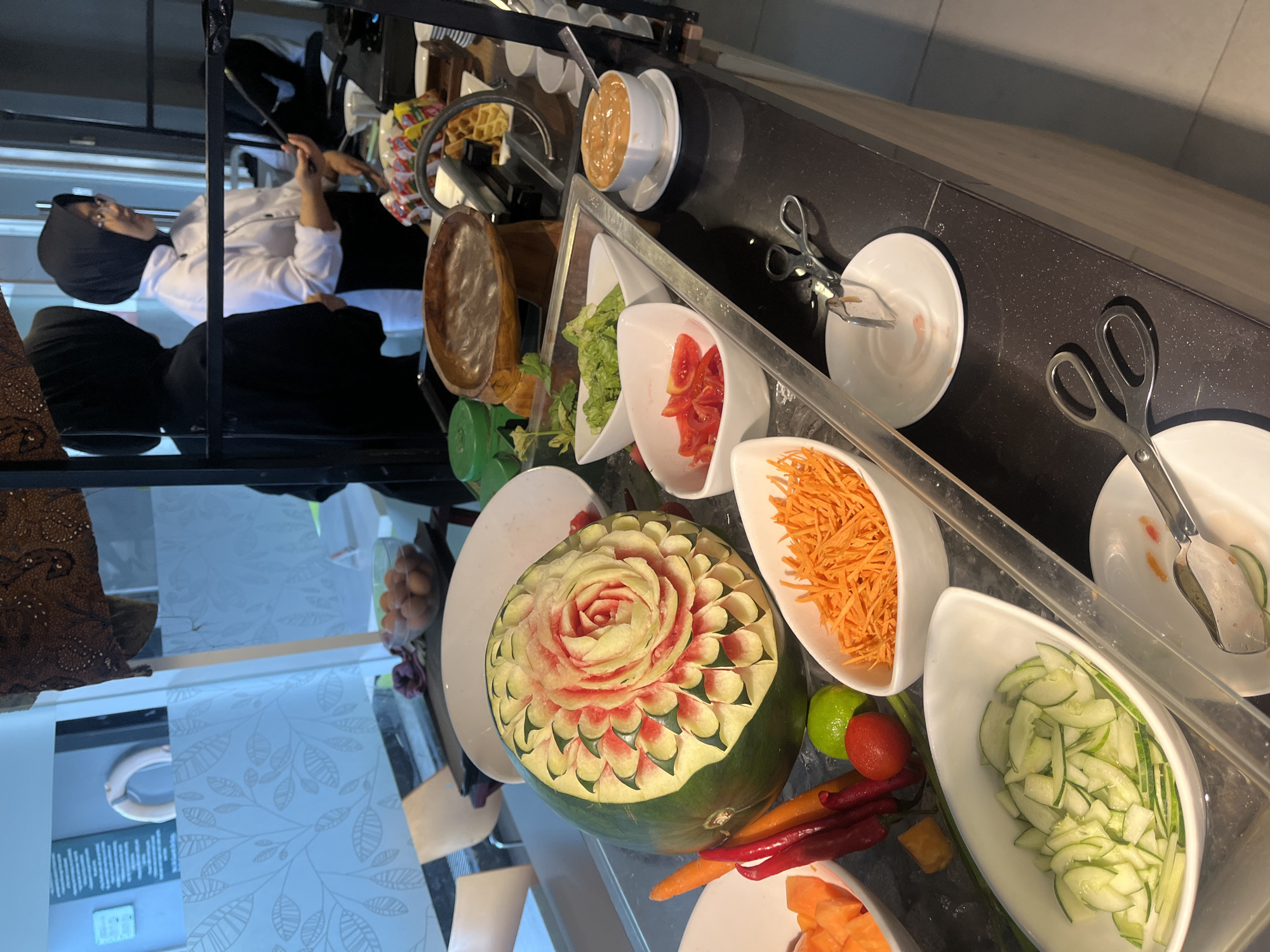The image shows a buffet station featuring a variety of fresh ingredients. In the foreground, there are bowls of sliced cucumbers, shredded carrots, and chopped tomatoes. A whole watermelon carved into a decorative flower shape is prominently displayed. Other items visible include chili peppers, carrots, a lime, and a bowl of what appears to be a sauce or dip. In the background, two women in chef attire are visible. One is wearing a black hijab and the other a white chef coat and black hijab, with glasses. They appear to be serving or preparing food. The buffet is set up indoors, with a reflective surface on the counter. The overall impression is that of a food service area, possibly at a hotel or restaurant, in Gapurosukolilo, Indonesia.