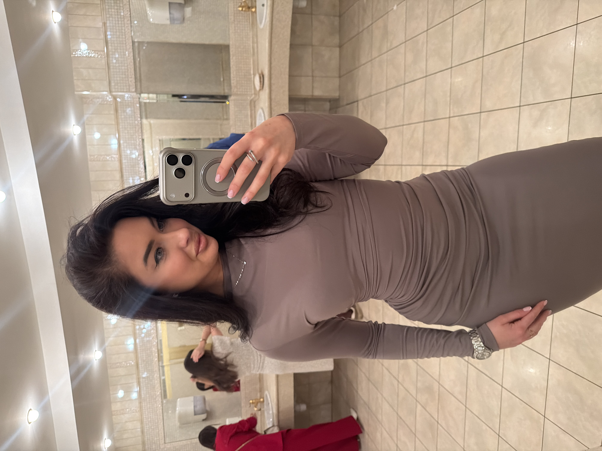 The image presents a mirror selfie taken by a young woman in what appears to be a public restroom or powder room in Moscow, Russia. The main subject, a woman with long dark hair, light eyes, and a pleasant expression, is positioned towards the left side of the frame, looking at the camera with a slight smile. She is wearing a form-fitting, long-sleeved, high-neck dress in a neutral taupe or light brown shade, which features gathering or ruching on the side. She accessorizes with a delicate silver necklace, a ring on her left ring finger, and a silver-toned watch on her left wrist. Her nails are neatly manicured with a French tip design. She holds a light grey smartphone, equipped with a triple camera system and a prominent MagSafe ring on its back, to capture her reflection.
The setting is an interior space characterized by modern amenities. The walls are covered with light-colored rectangular tiles, and large mirrors with decorative, possibly sparkling, borders line the walls. Bright, warm artificial lighting emanates from several wall-mounted sconces, creating reflections in the mirrors. The floor is tiled with larger, square, light-colored tiles.
In the background, visible through the mirror's reflection, are glimpses of other people. One individual with long dark hair, wearing a light-colored top, is seen from the back standing at a sink. Another person, also seen from behind, is dressed in a vibrant red outfit, further in the room near another sink area. The overall impression is that of a well-maintained, possibly upscale, public facility where multiple individuals are present. No discernible text is visible within the image.