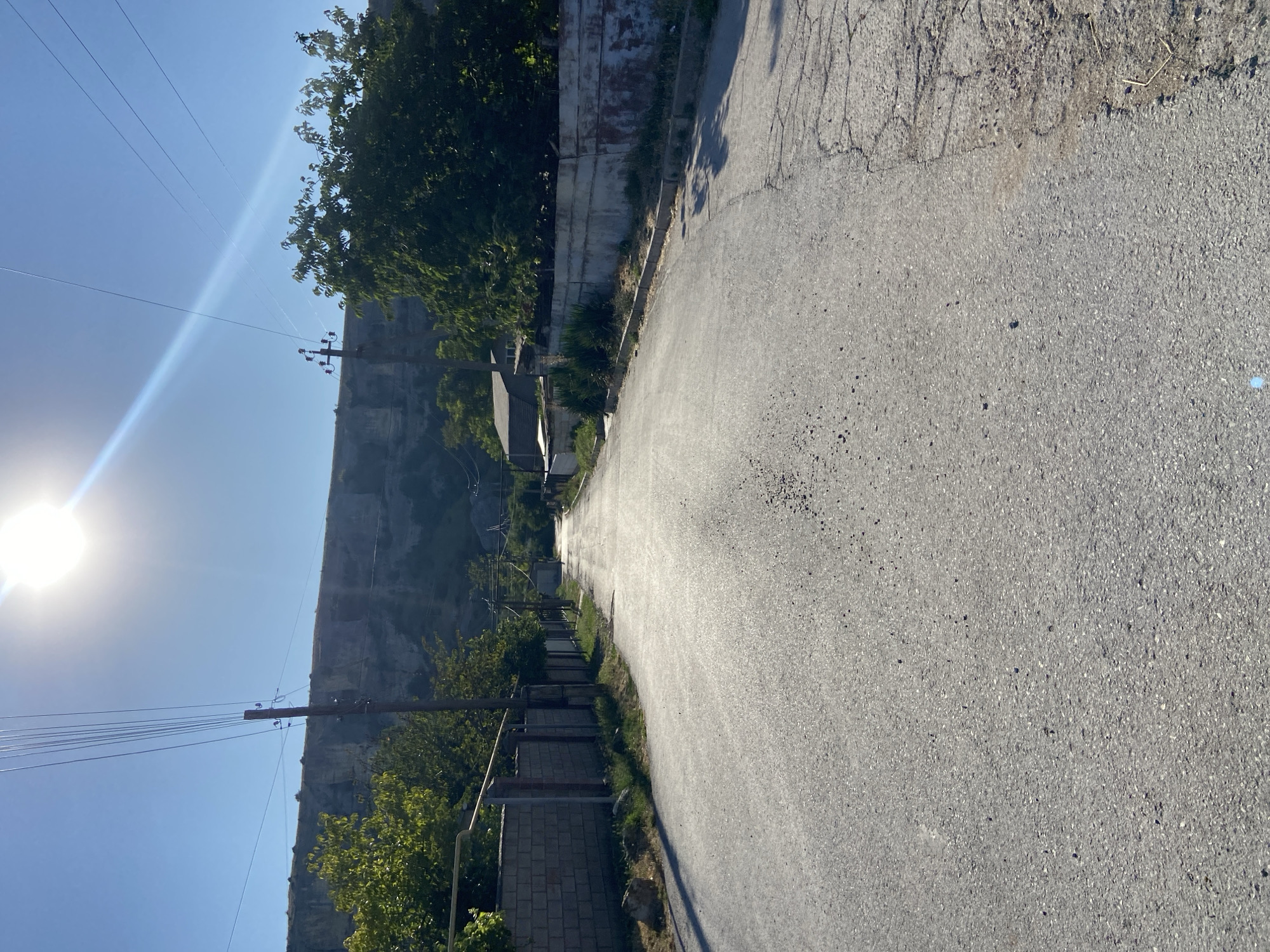 A straight, paved road extends into the distance, lined on both sides by low walls and vegetation. On the left, a cinder block wall is visible, while the right side features a weathered concrete wall with some graffiti. Overhead, power lines stretch across the clear blue sky, with the sun prominently displayed and creating lens flare. In the background, a steep, rocky cliff face rises above the trees and houses that dot the landscape. The road appears to be in a rural or semi-rural area, possibly a village. The overall impression is one of a bright, sunny day.