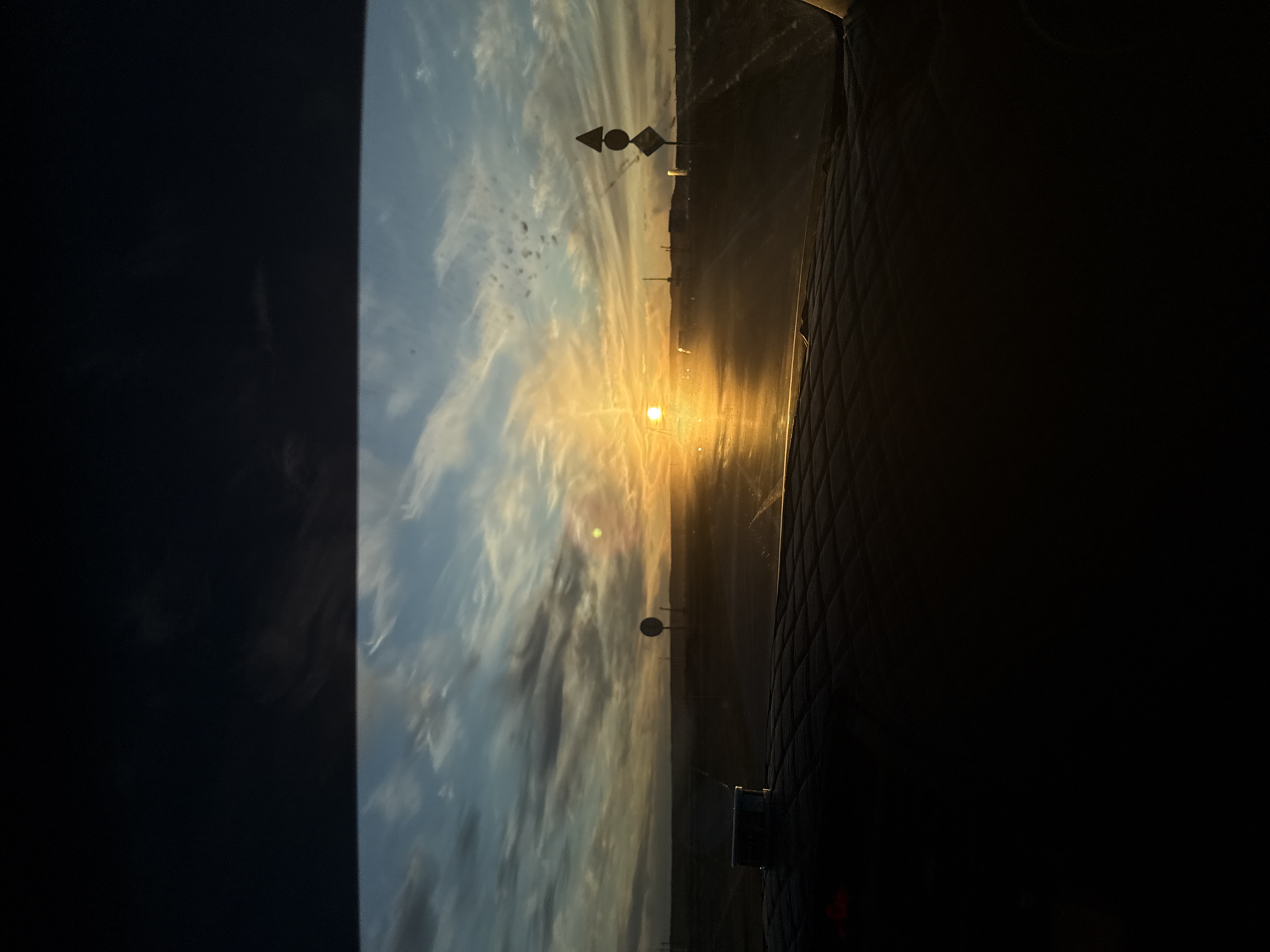 The image captures a view through a window or windshield at sunset. The sky is filled with clouds streaked with light from the setting sun, which is the brightest point of the image. There are several utility poles and what may be road signs in the distance. The foreground of the image, particularly the right side, is dark and features what appears to be a quilted surface, possibly part of a car interior or a padded wall. There's no visible sign of Tulagino, Russia.