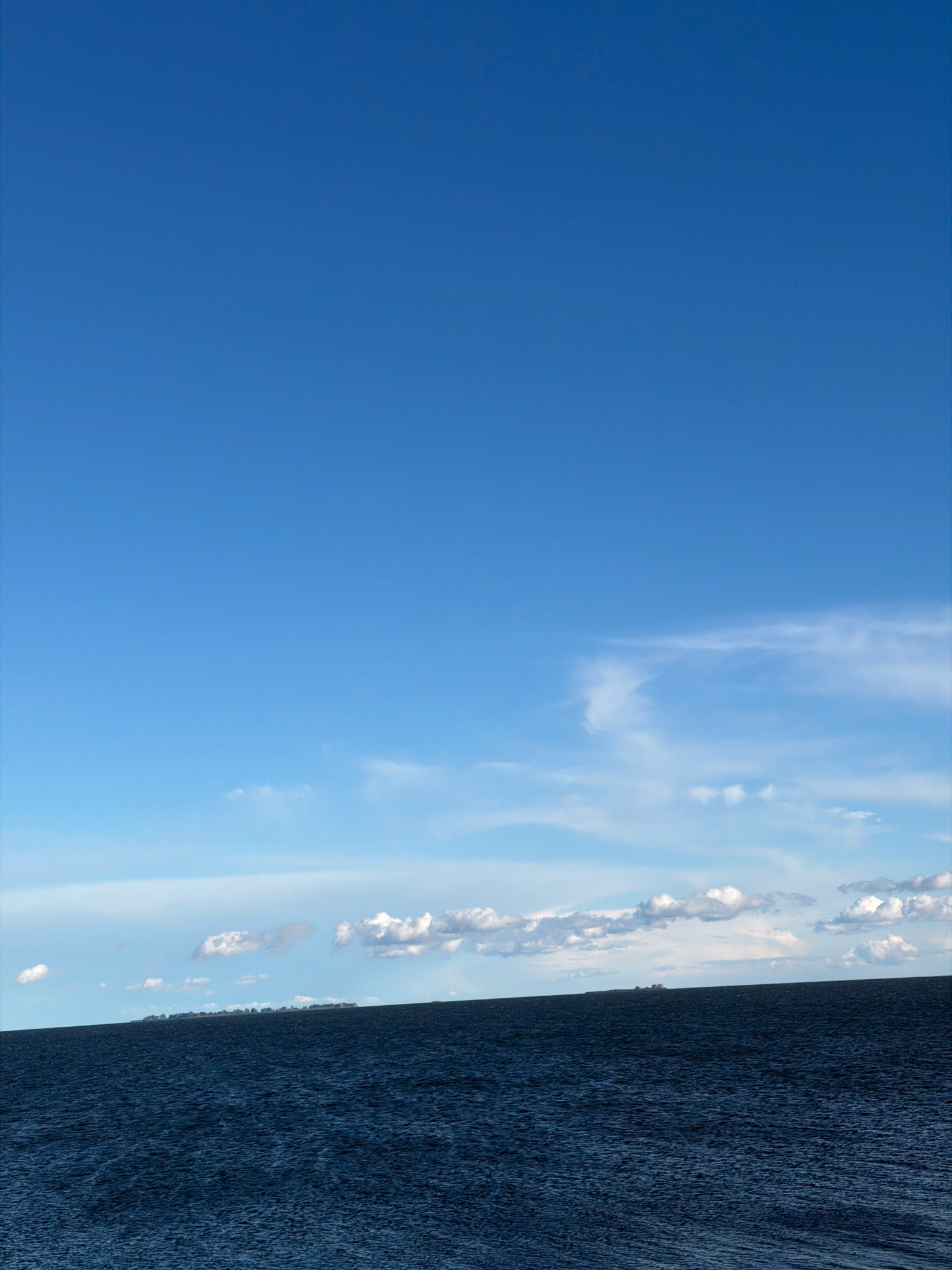 A wide shot captures a vast expanse of dark blue water under a clear blue sky. The water has a textured surface, suggesting gentle waves. A distant, low-lying landmass is visible on the horizon, dotted with trees or buildings. Scattered white clouds drift in the sky, some appearing wispy and others more puffy. The image is devoid of any human presence or discernible urban or natural landmarks that would indicate a specific location within a city.