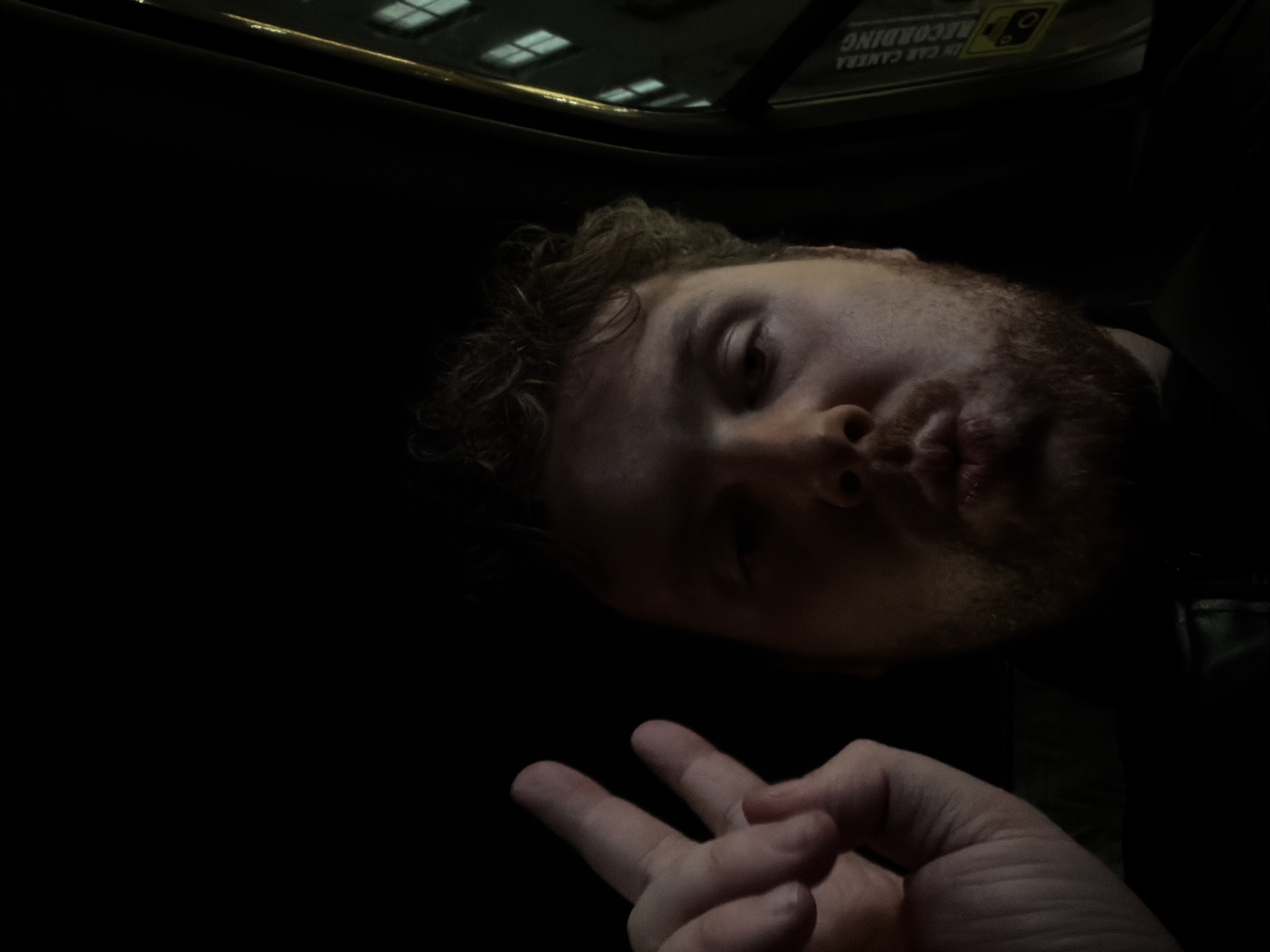 The image appears to be taken inside a vehicle, possibly a taxi, in New York City. A man is visible, angled to the side, with a reddish beard and curly hair. He is making a peace sign with his fingers. The background is dark, with a partial view of buildings visible through the vehicle's roof. A sign that reads "IN CAR CAMERA RECORDING" is also visible.