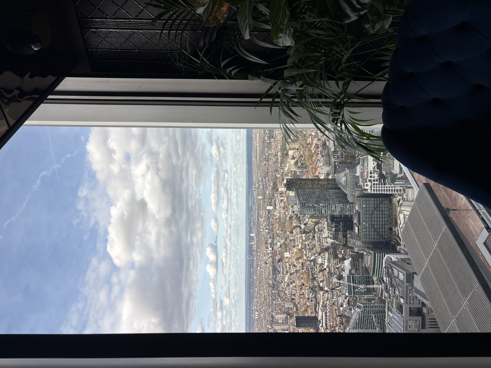 From a high vantage point, a view of London, United Kingdom is visible through a window. The sky is partly cloudy. A city skyline includes buildings of various heights and styles. A plant and a blue tufted chair are also visible.