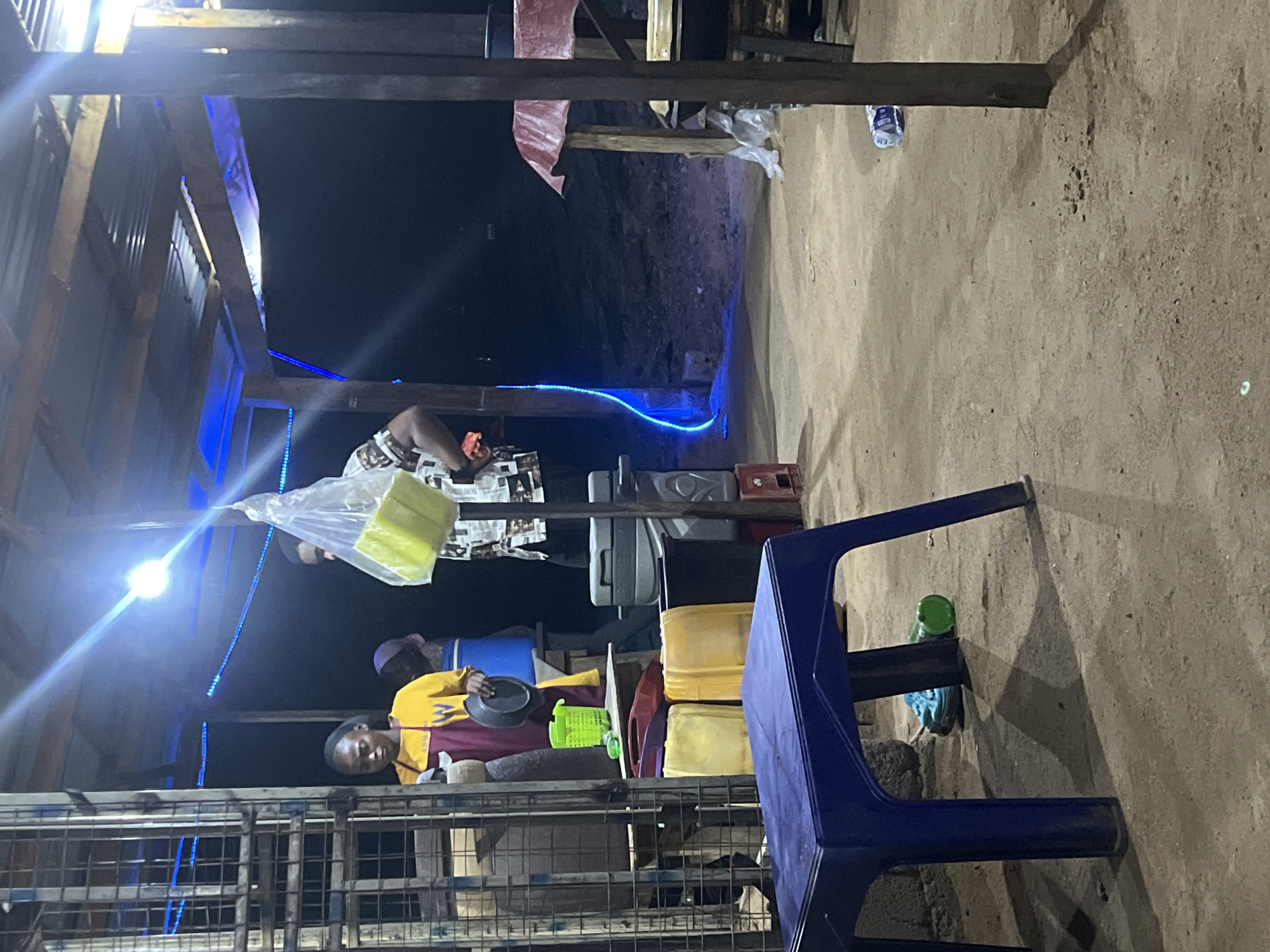 The image depicts an outdoor scene, likely a small business or market stall, in Jalingo, Nigeria. Two people are visible inside the stall. One person is wearing a yellow and maroon shirt and is holding a dark-colored plate or bowl. The other individual is partially visible and appears to be handling goods wrapped in newspaper and plastic. The stall itself is constructed with a metal grid and wooden beams, and is illuminated by blue lights, including a string of LED lights. Yellow containers are visible on the shelves. A blue plastic chair sits outside the stall. The ground is dirt.