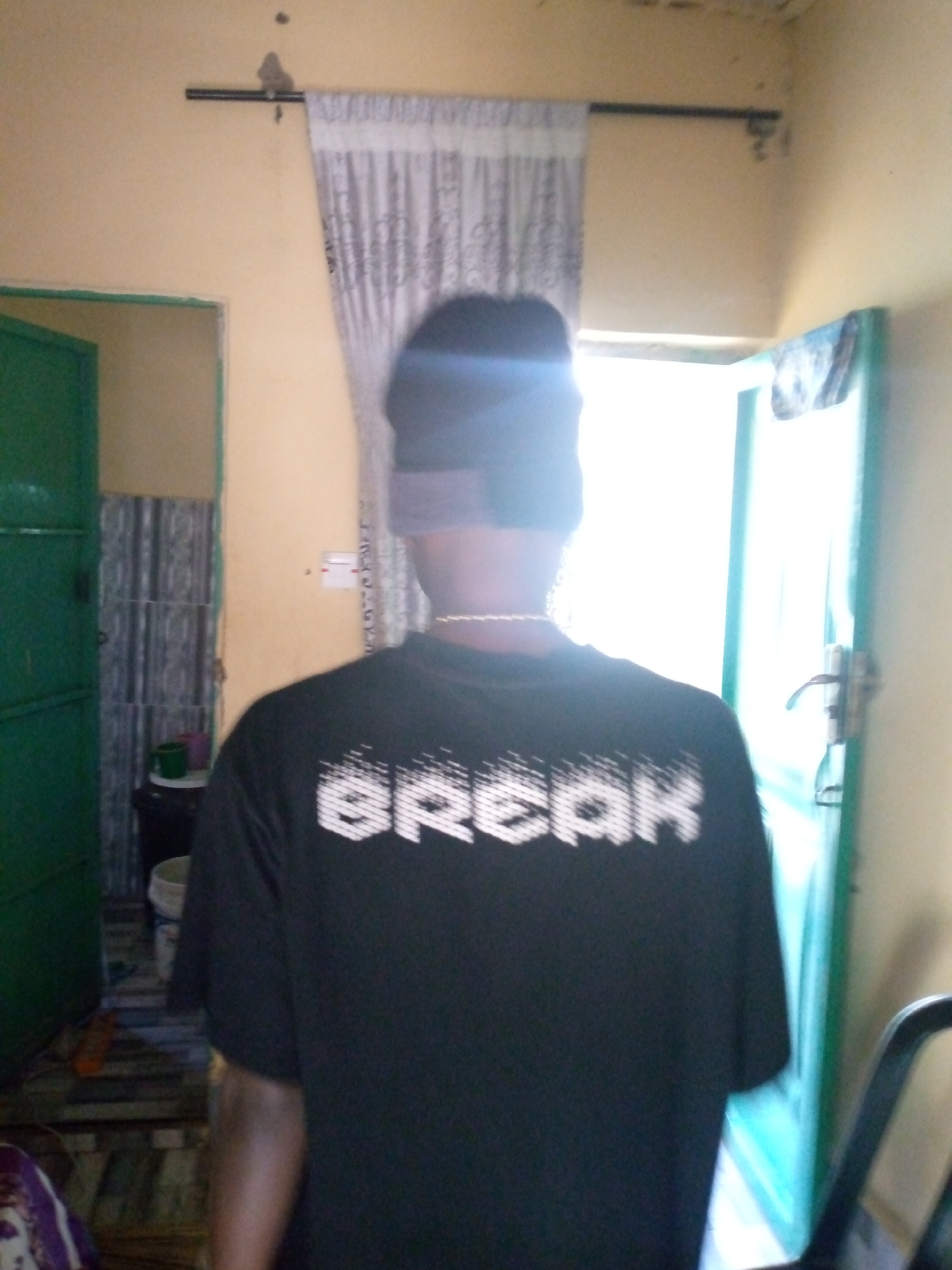 The image depicts the back of a person wearing a black t-shirt with the word "BREAK" printed in white, stylized block lettering across the back. The person is also wearing a dark head covering that obscures their hair and forehead. A thin, gold-colored chain is visible around their neck. The person appears to be indoors, in what looks like a domestic setting in Jalingo, Nigeria. To the left of the person, a green door is visible, slightly ajar. To the right, another green door, this one seemingly leading outdoors or to another room, is also visible, with a handle on it. Between the two doors, a window or opening is brightly lit by natural light. The walls are painted a pale yellow. A curtain with a subtle pattern hangs near the top of the frame. In the background, on the left, a tiled wall is partially visible. A bucket and possibly other objects are situated on the floor near the green door on the left. The overall scene is casually arranged, suggesting an everyday indoor environment.