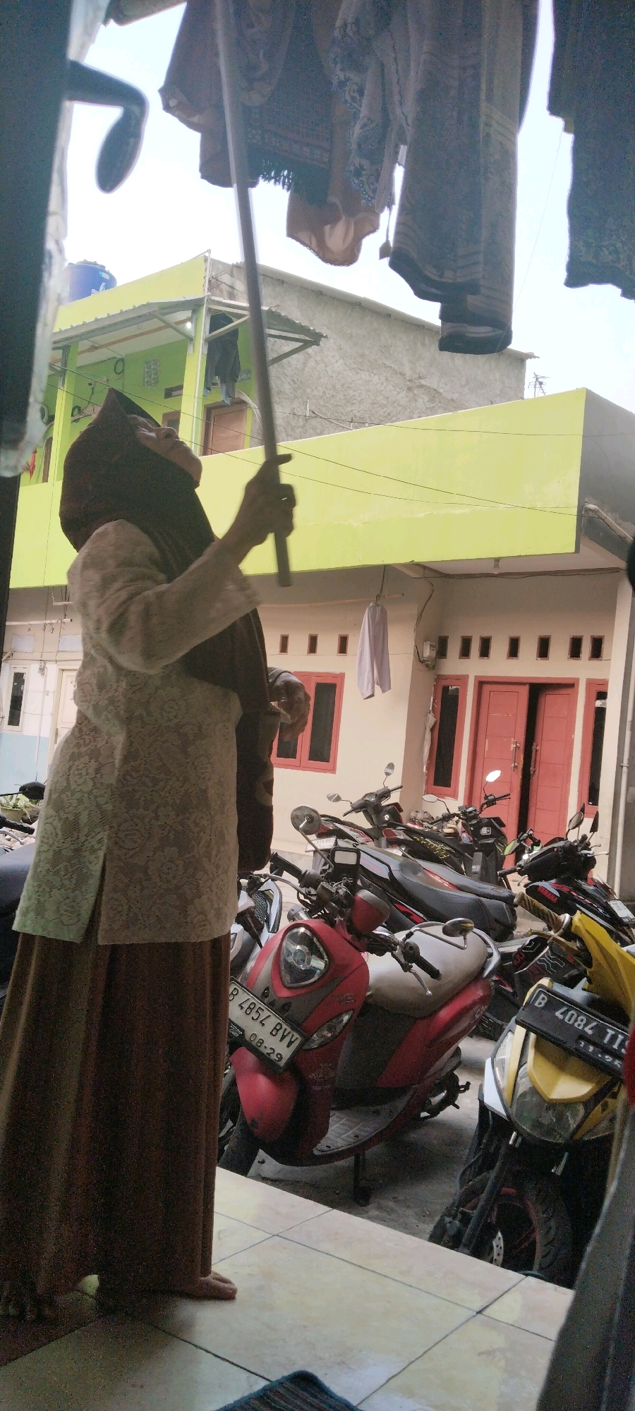 A woman wearing a brown hijab and a patterned white blouse with a long brown skirt stands in what appears to be a residential area in West Jakarta, Indonesia. She is holding a long pole and looking upwards, potentially interacting with laundry hanging overhead, which is partially visible in the foreground. In the background, a light yellow building with red-framed windows and doors is visible. A row of parked motorcycles, including a red scooter with the license plate B 4854 BVV, are lined up in front of the building. The ground is tiled, and the overall scene suggests a domestic setting, possibly a backyard or alleyway.